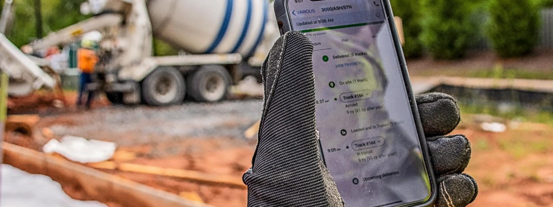 Smartphone with open app is held by a gloved hand; a ready-mix truck is visible in the background