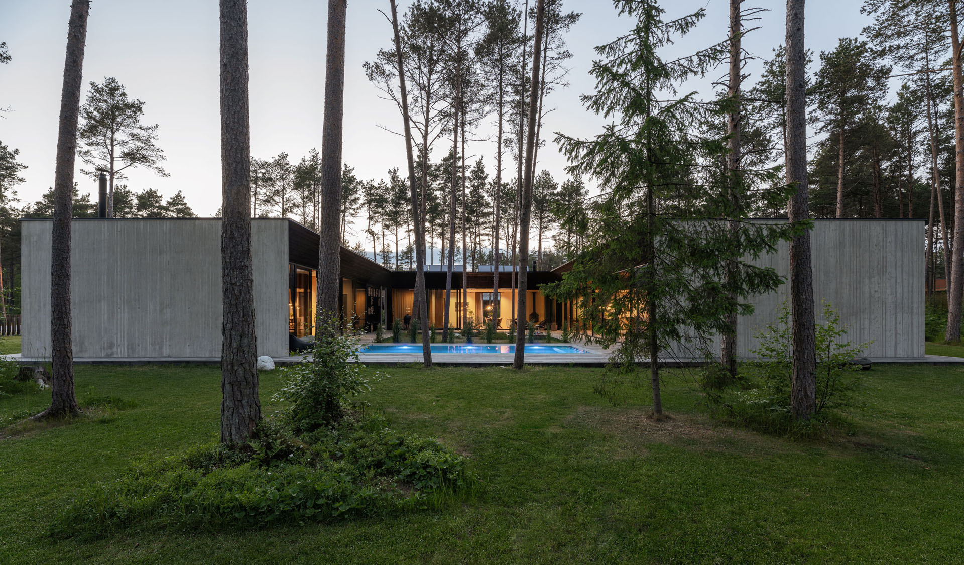 Bungalow made of exposed concrete, surrounded by individual trees, with an illuminated pool in the inner courtyard