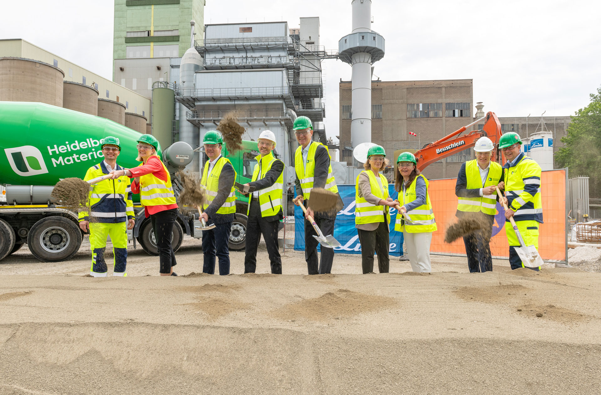 Plant site with 9 people in protective clothing and shovels in hand in front of a pile of sand