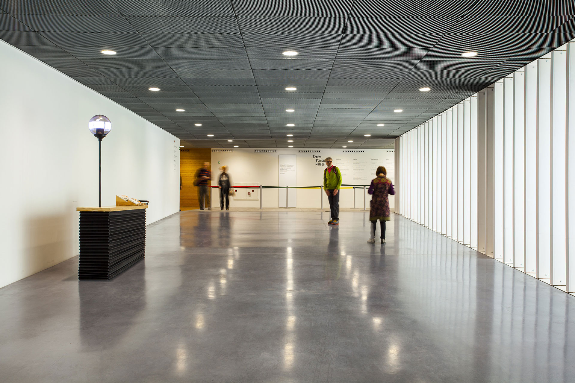 View into a wide building corridor with a reflective concrete floor and symmetrically arranged lamps in the ceiling. Four people are standing in the corridor, on the left there is a counter with a light.