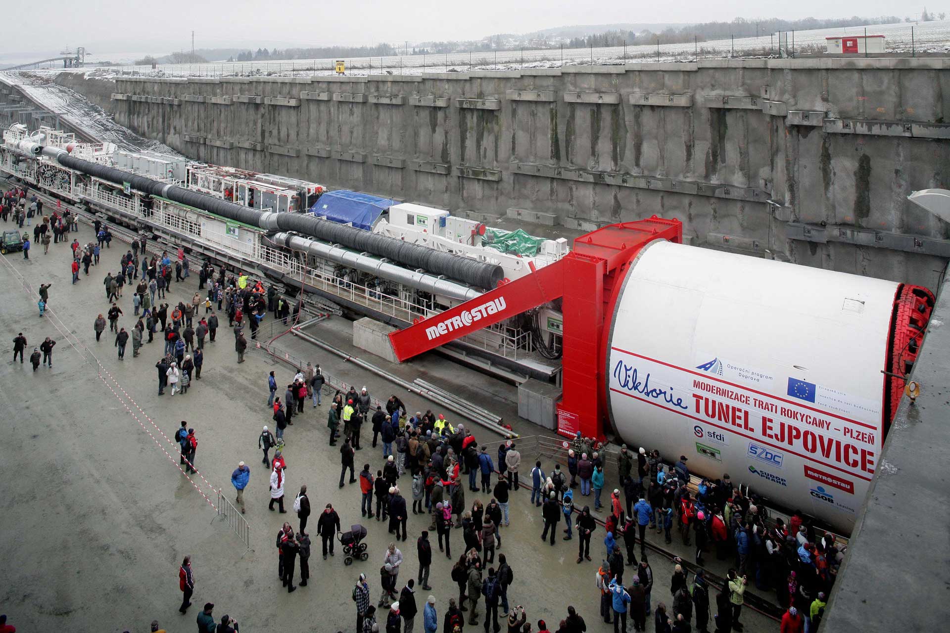 Ein großer Tunnelbohrer mit rot-weißer Außenhülle und der Aufschrift “Metrostav” sowie “Tunnel Excavation - EPB Shield” steht auf einer Betonfläche, bereit für den Beginn eines Tunnelbauprojekts, mit einer langen Struktur, die in die Ferne reicht.