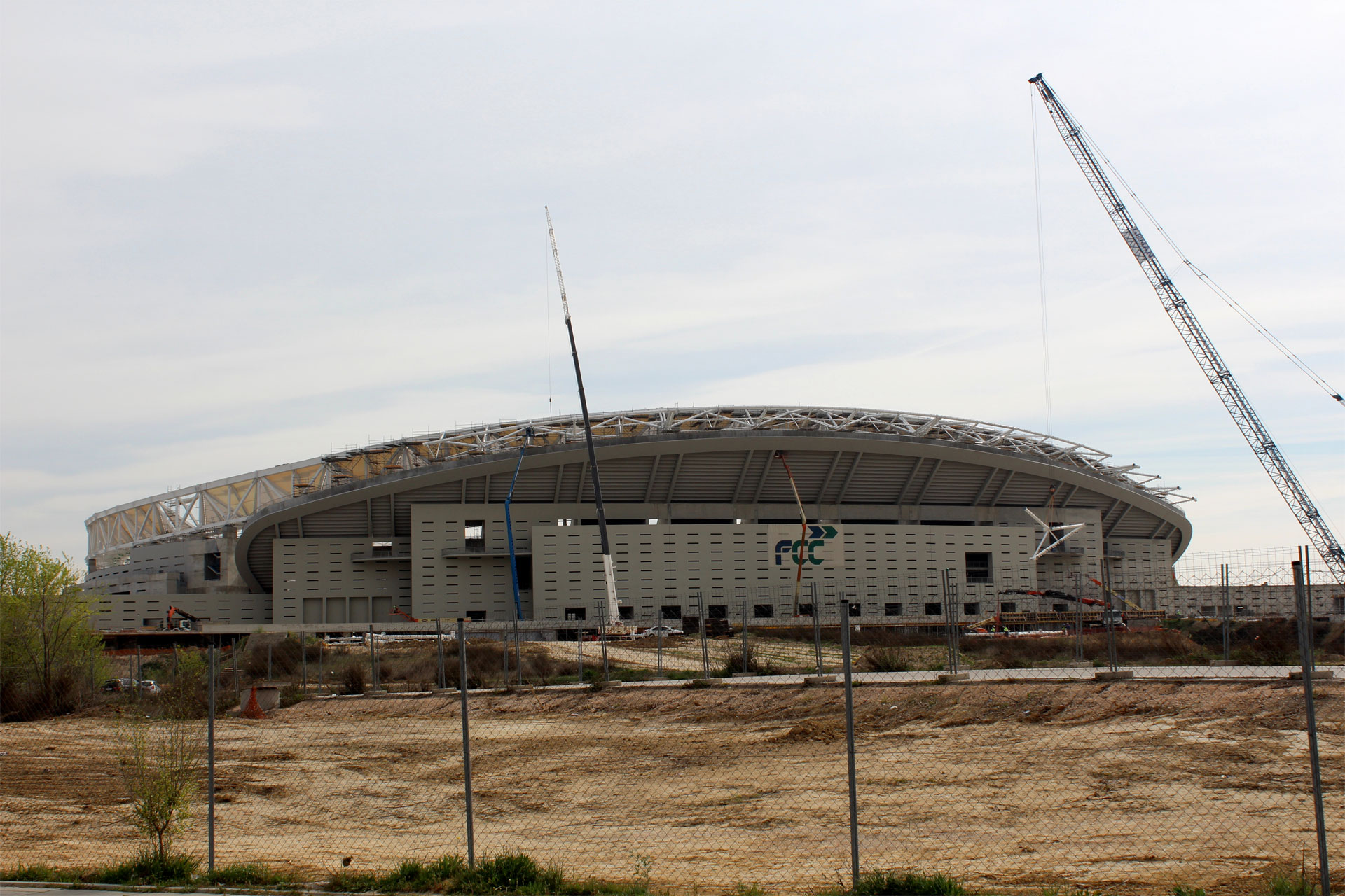 An under-construction stadium with scaffolding and an incomplete roof, a crane to the right, and a barren field leading to it