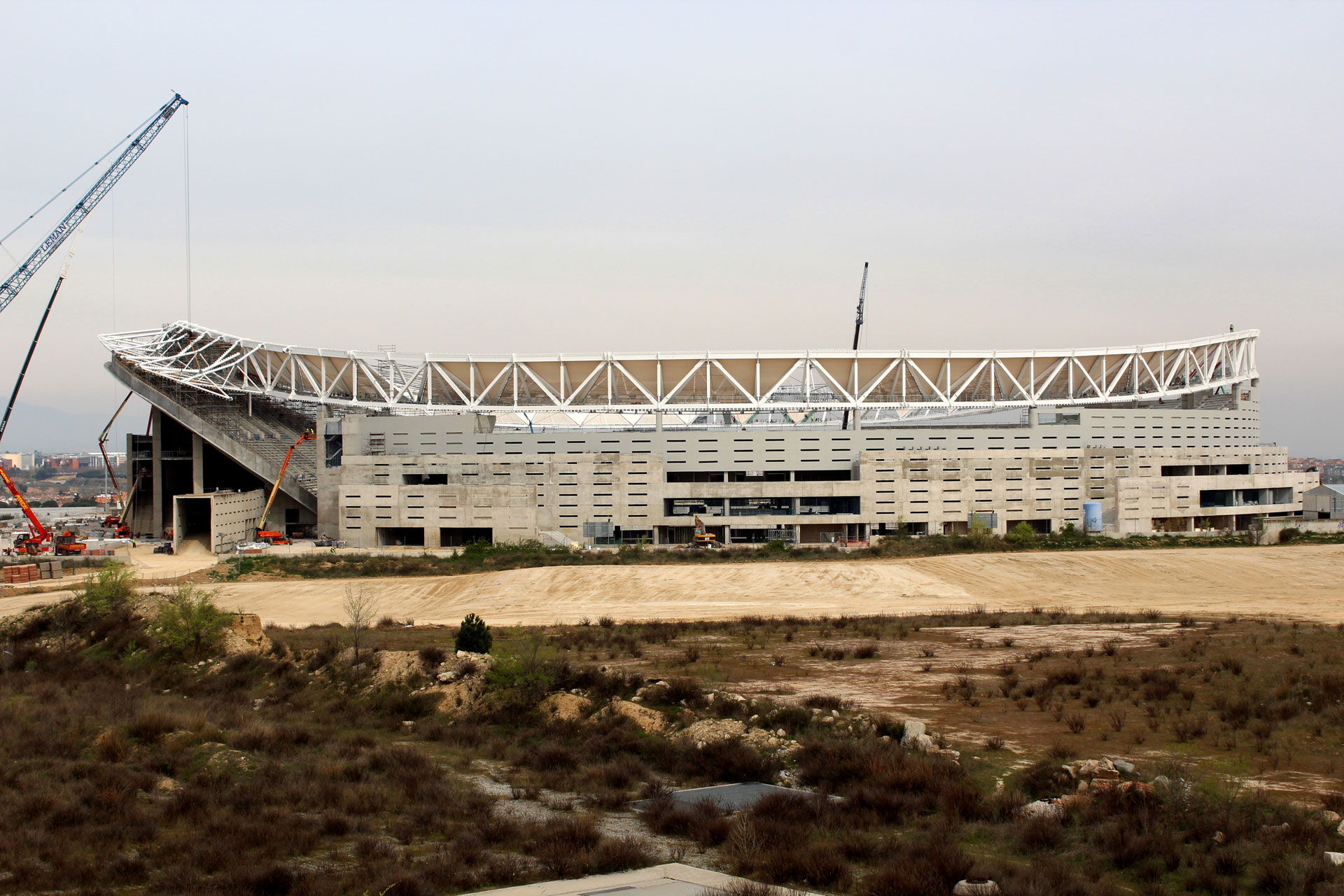 Ein teilweise fertiggestelltes Stadion mit freiliegendem Gerüst und Kränen vor einem bewölkten Himmel