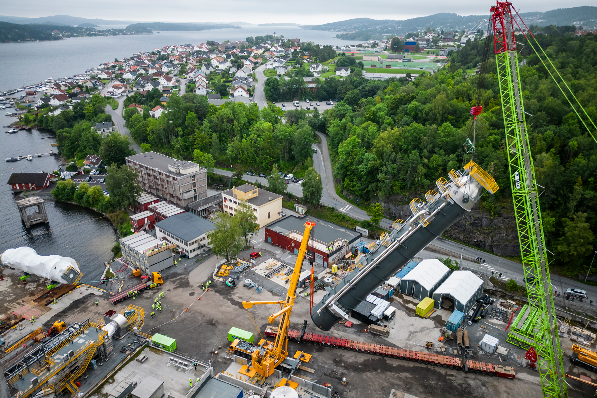 Aerial view of a cement plant with a large crane near a body of water, with residential areas in the background.