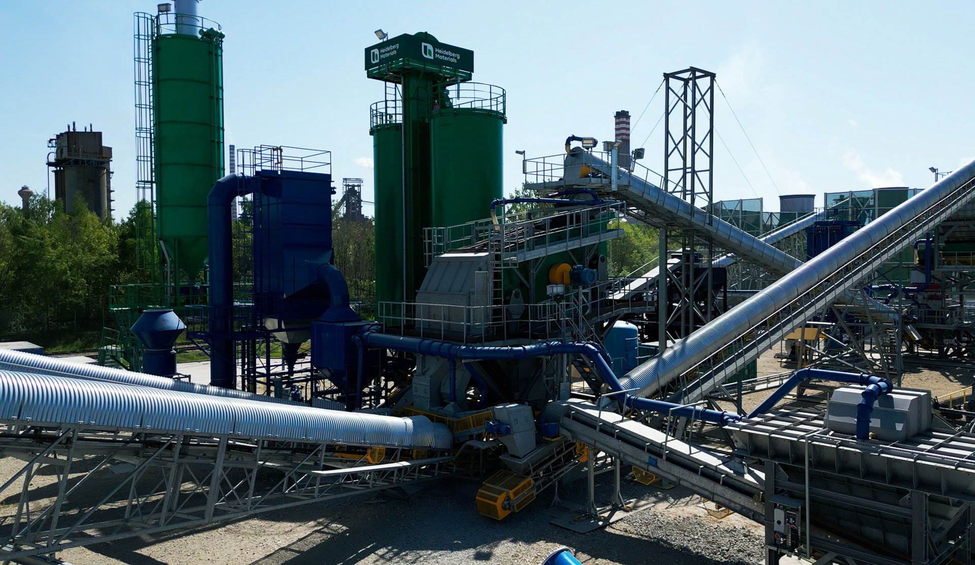 Ein Industriekomplex mit großen zylindrischen Lagertanks in Grün- und Blautönen, umgeben von einem komplexen Netzwerk aus Metallrohren und -strukturen unter einem klaren Himmel.