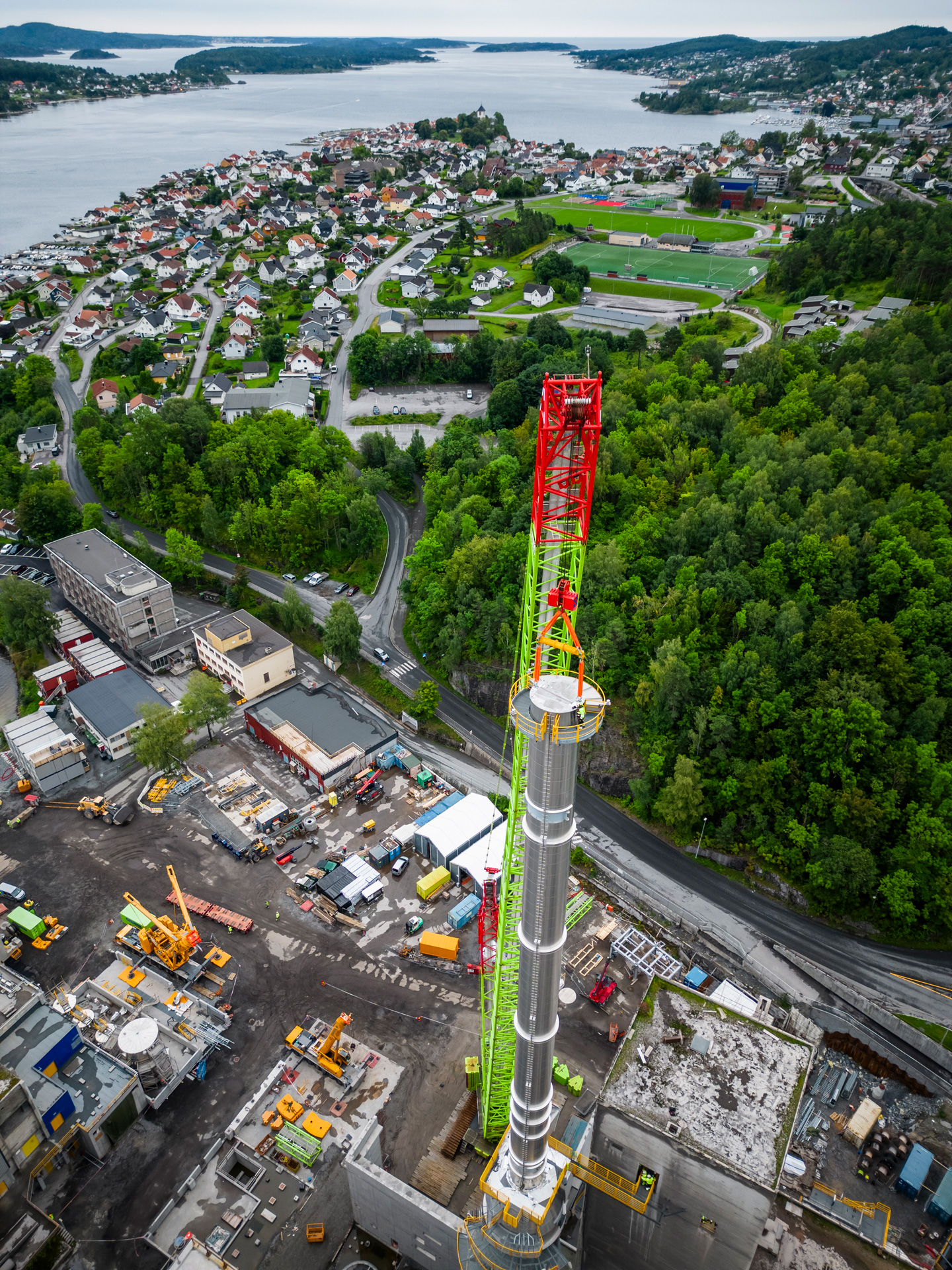 Aerial view of a construction crane towering over a cement plant near a residential area with lush greenery and a body of water in the background.