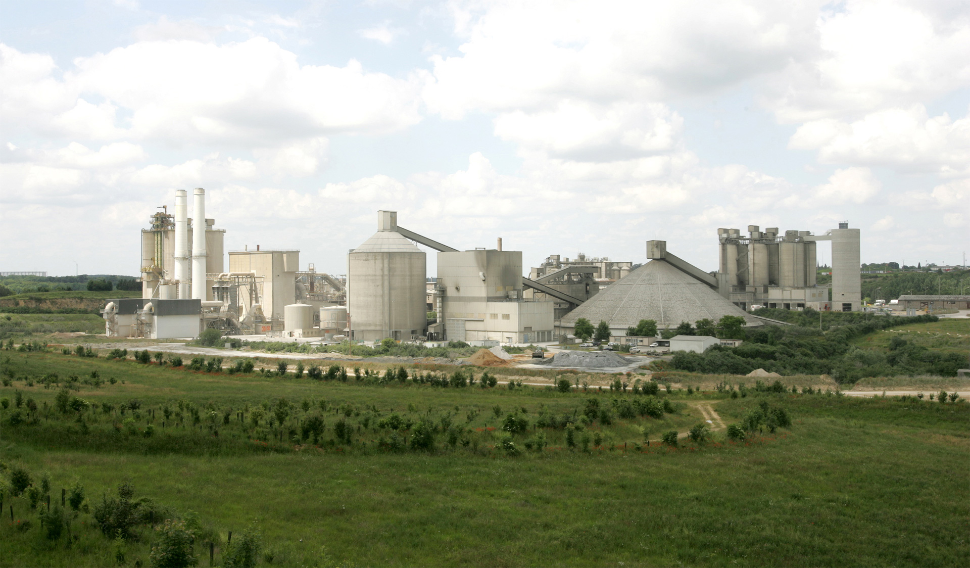 Industrial cement plant with silos amidst green fields under a cloudy sky