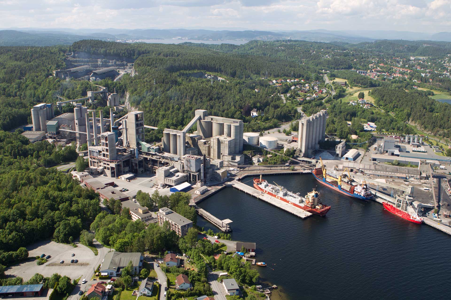 An aerial view of a large industrial facility by the water, showcasing silos, tall buildings, and a docked ship at a pier, surrounded by forest and distant buildings.