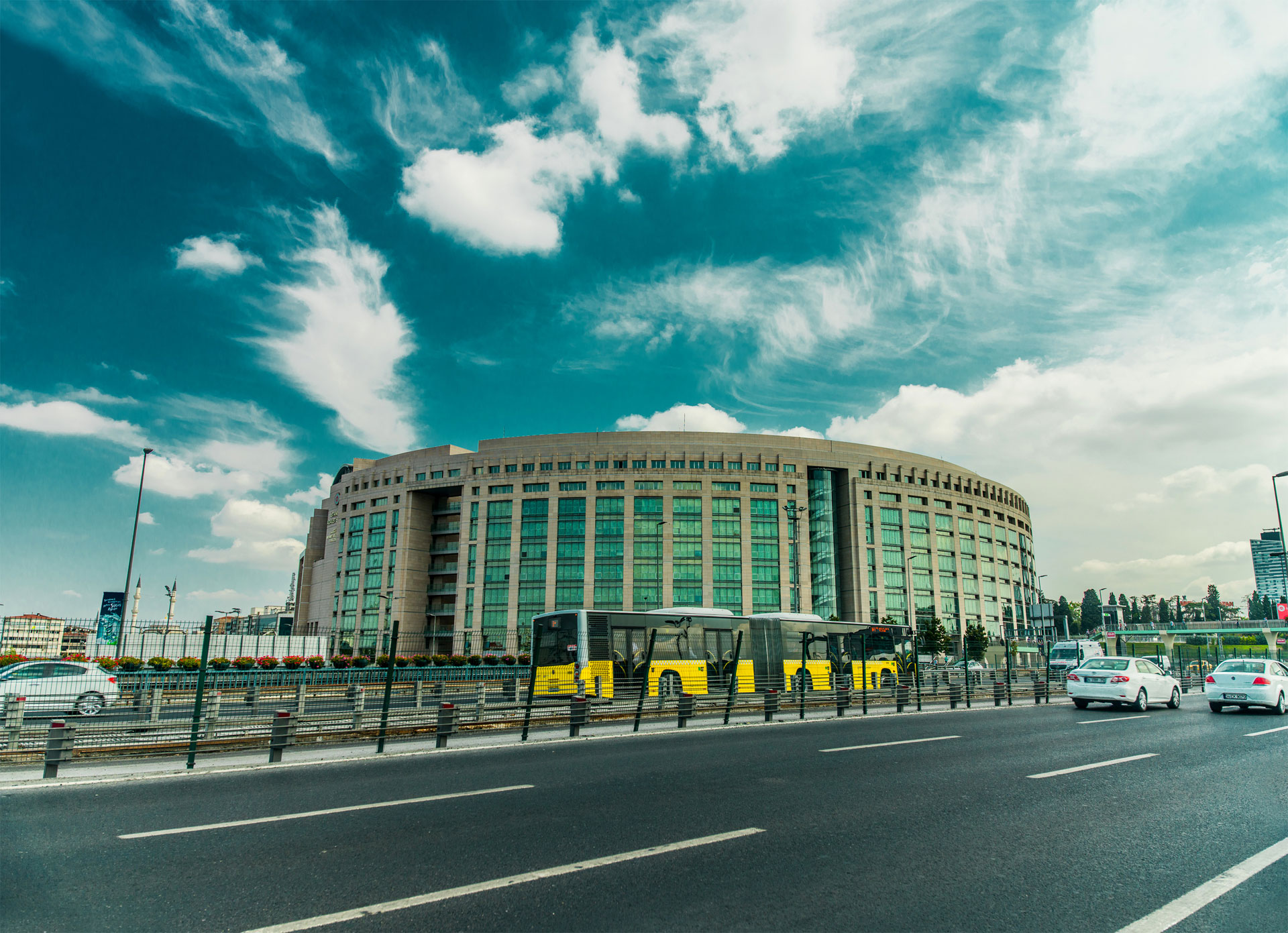 A modern circular building with a yellow tram passing by under a cloudy sky
