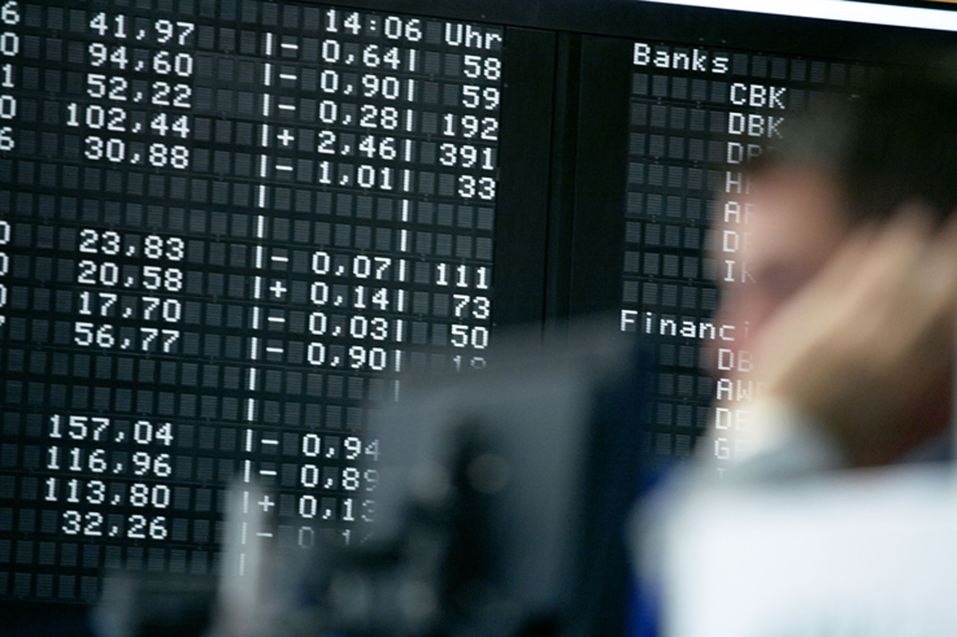 Eine verschwommene Figur im Vordergrund mit einer digitalen Börsentafel im Hintergrund, die verschiedene Zahlen und Aktiencodes anzeigt.