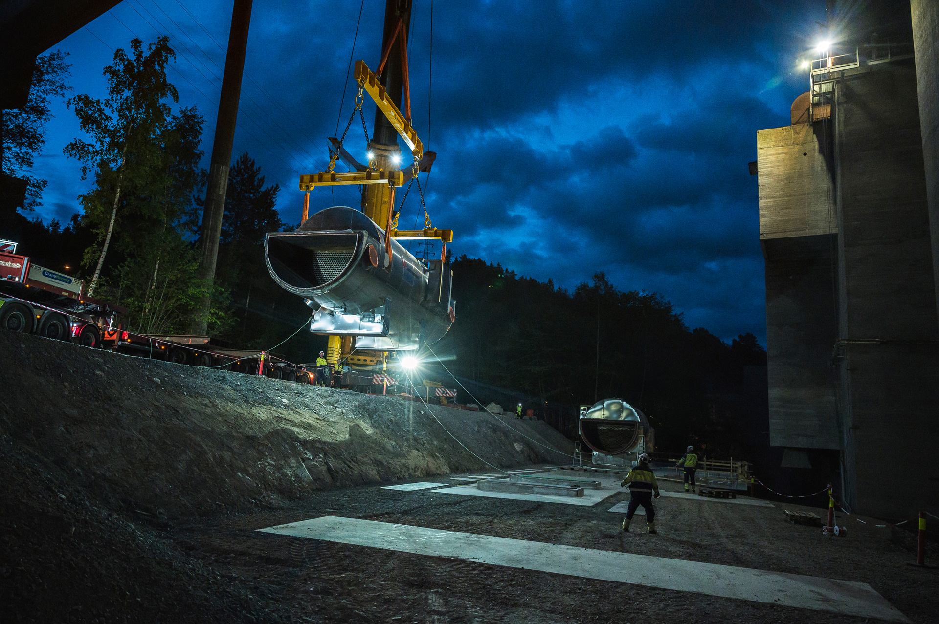 Construction site at twilight or night time with a large crane lifting a section of a CO₂ absorber. A similar sections is on the ground. Workers are present, and the scene is illuminated by artificial lighting.