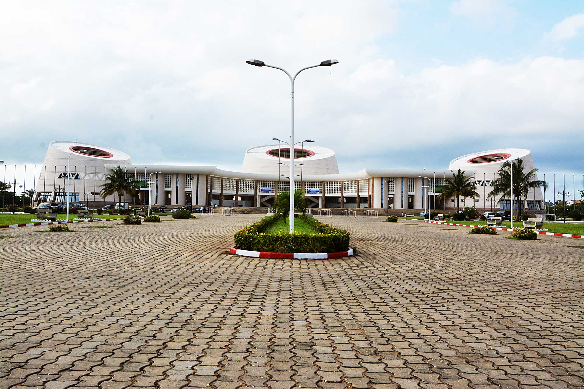 A large, modern building with a distinctive curved white facade and circular windows near the roof stands prominently. The foreground features a patterned pavement leading to the entrance, surrounded by street lamps and landscaped greenery under an overcast sky.