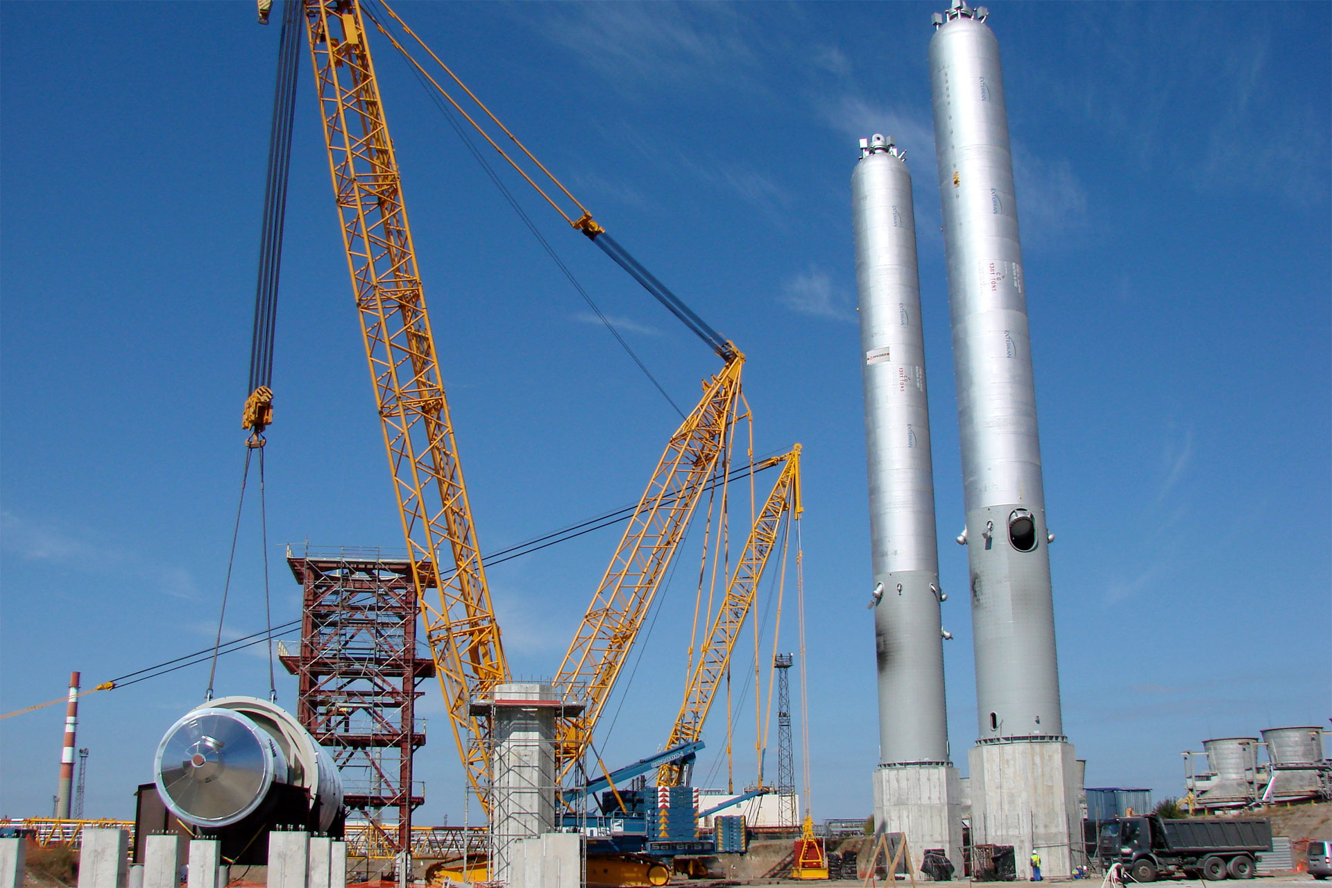 Large cranes lifting heavy cylindrical structures under a clear blue sky