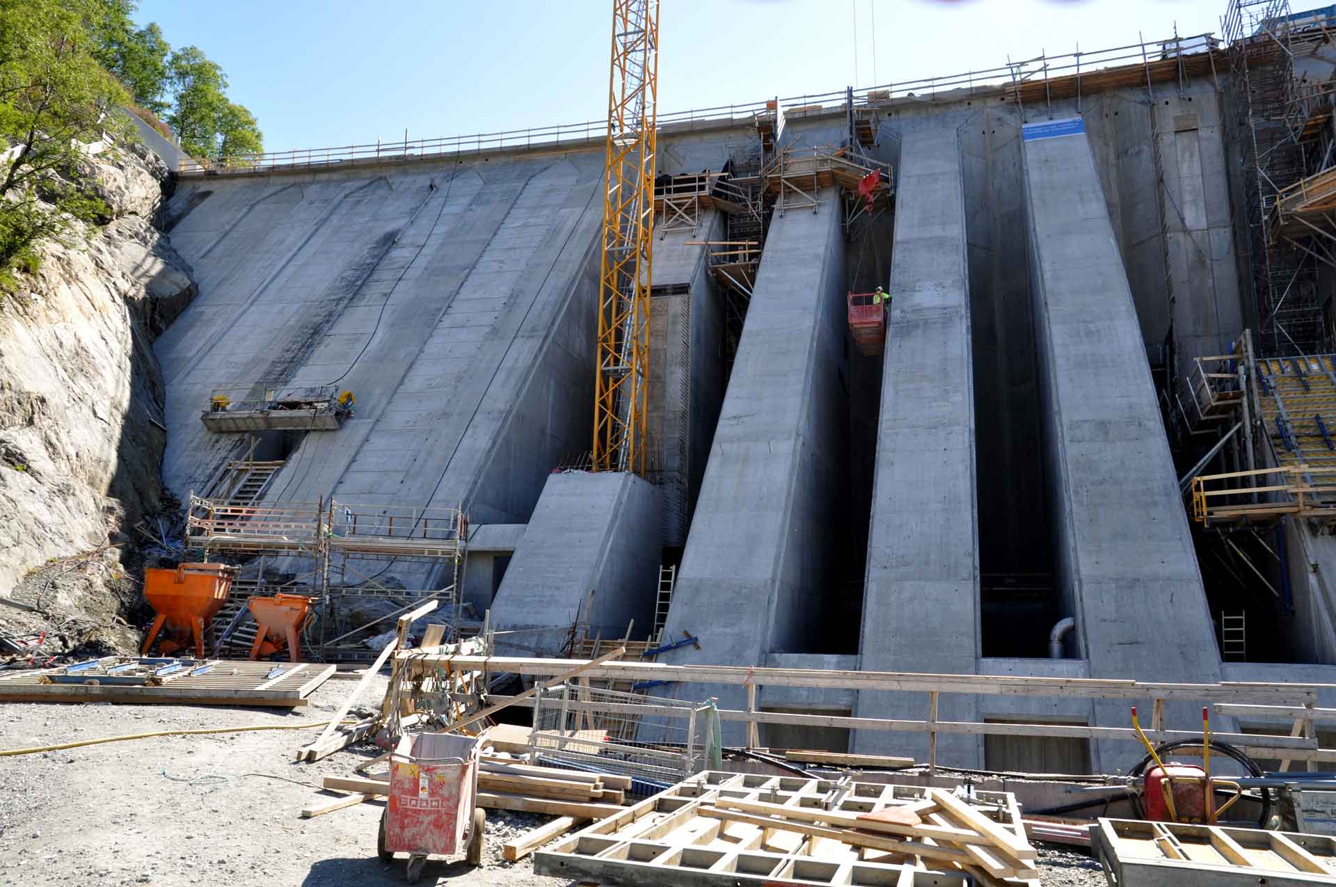 Eine große Betonstaumauer im Bau zeigt mehrere hohe, vertikale Betonwände mit Gerüsten und Baumaschinen, darunter Kräne und einen orangefarbenen Bagger am Fuß eines felsigen Abhangs.