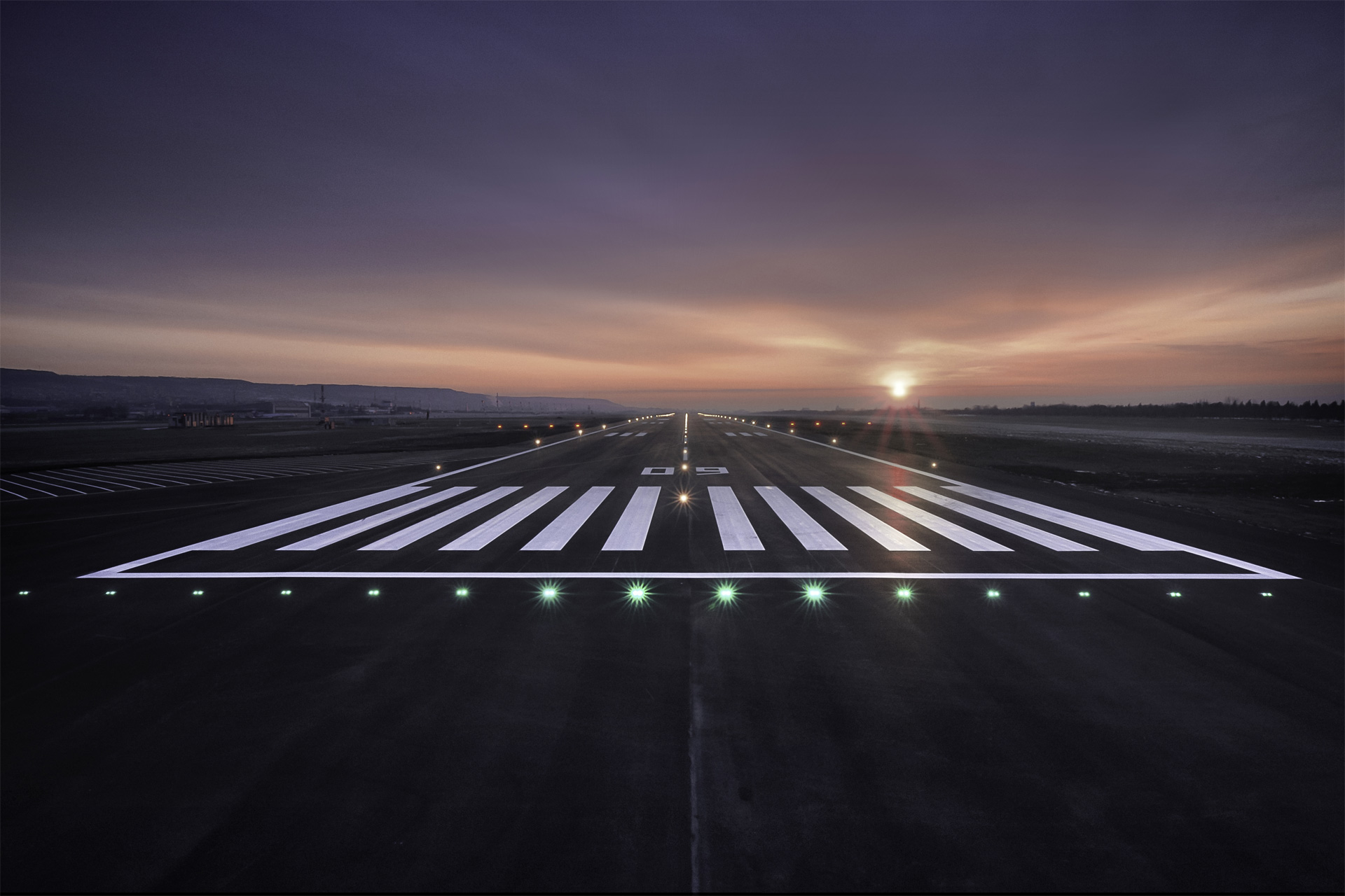 A runway at twilight with the sun near the horizon and green edge lights