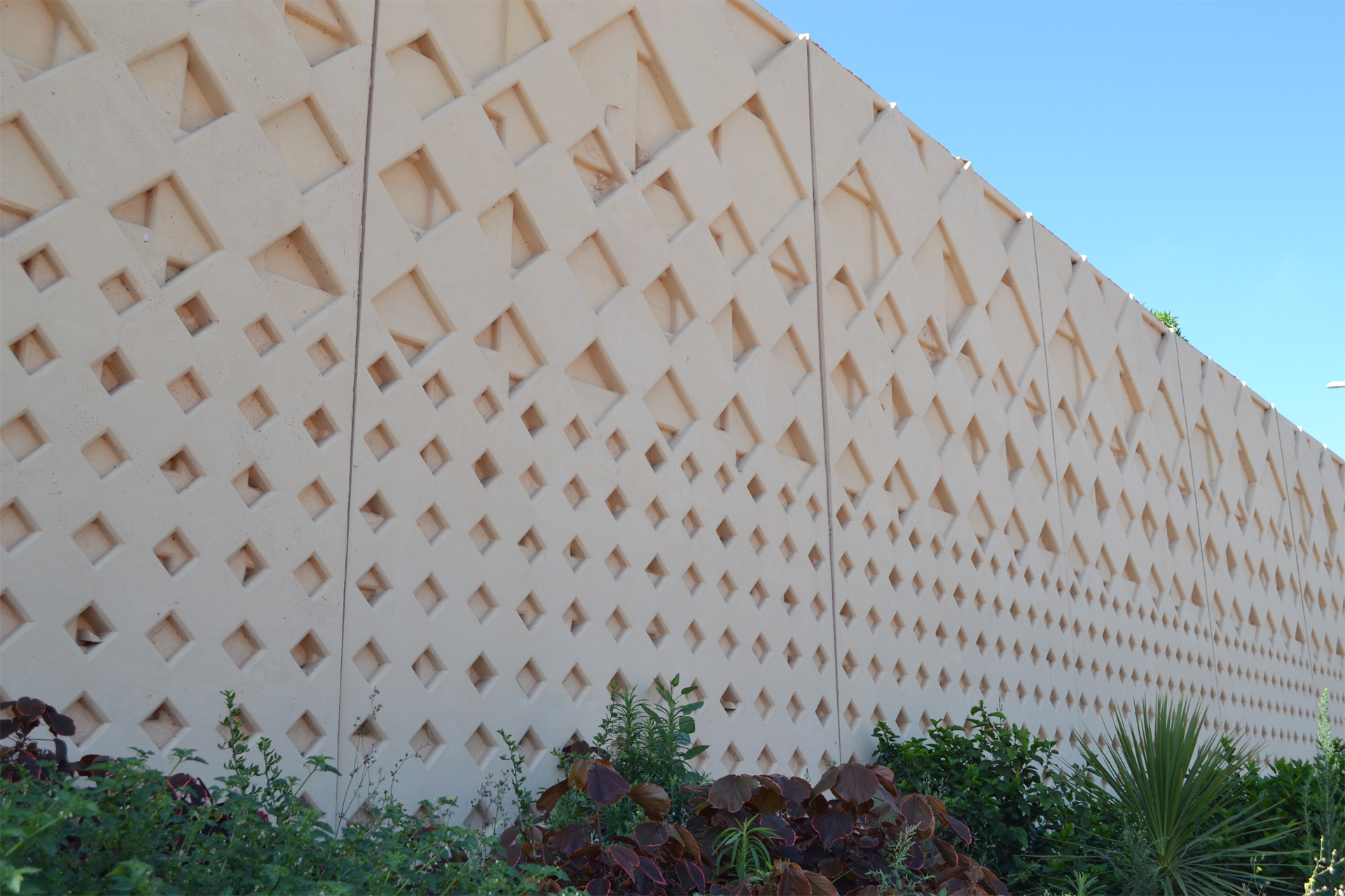 A wall with geometric diamond patterns and greenery at the base under a blue sky