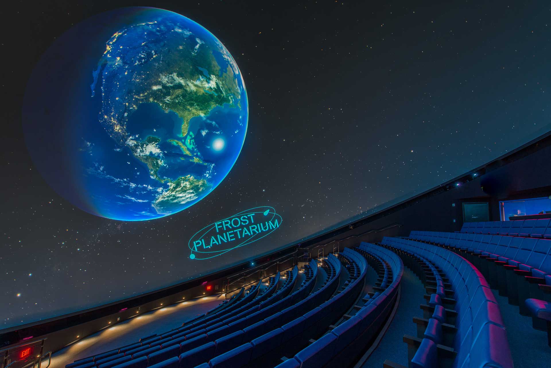 The interior of a planetarium with a detailed projection of Earth on the dome ceiling, seating arranged in concentric circles around the central display.