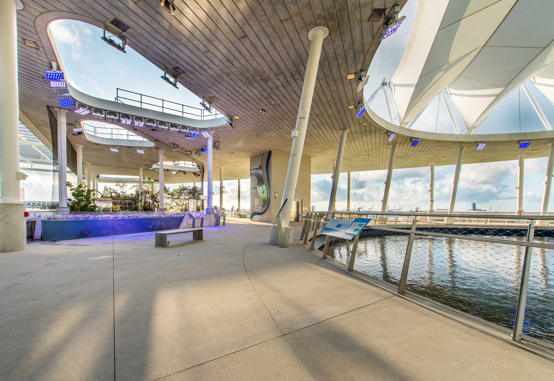  modern, covered pavilion with curved structures and support columns overlooking a water body. Benches and planters are present, and lighting fixtures are installed on the ceiling.