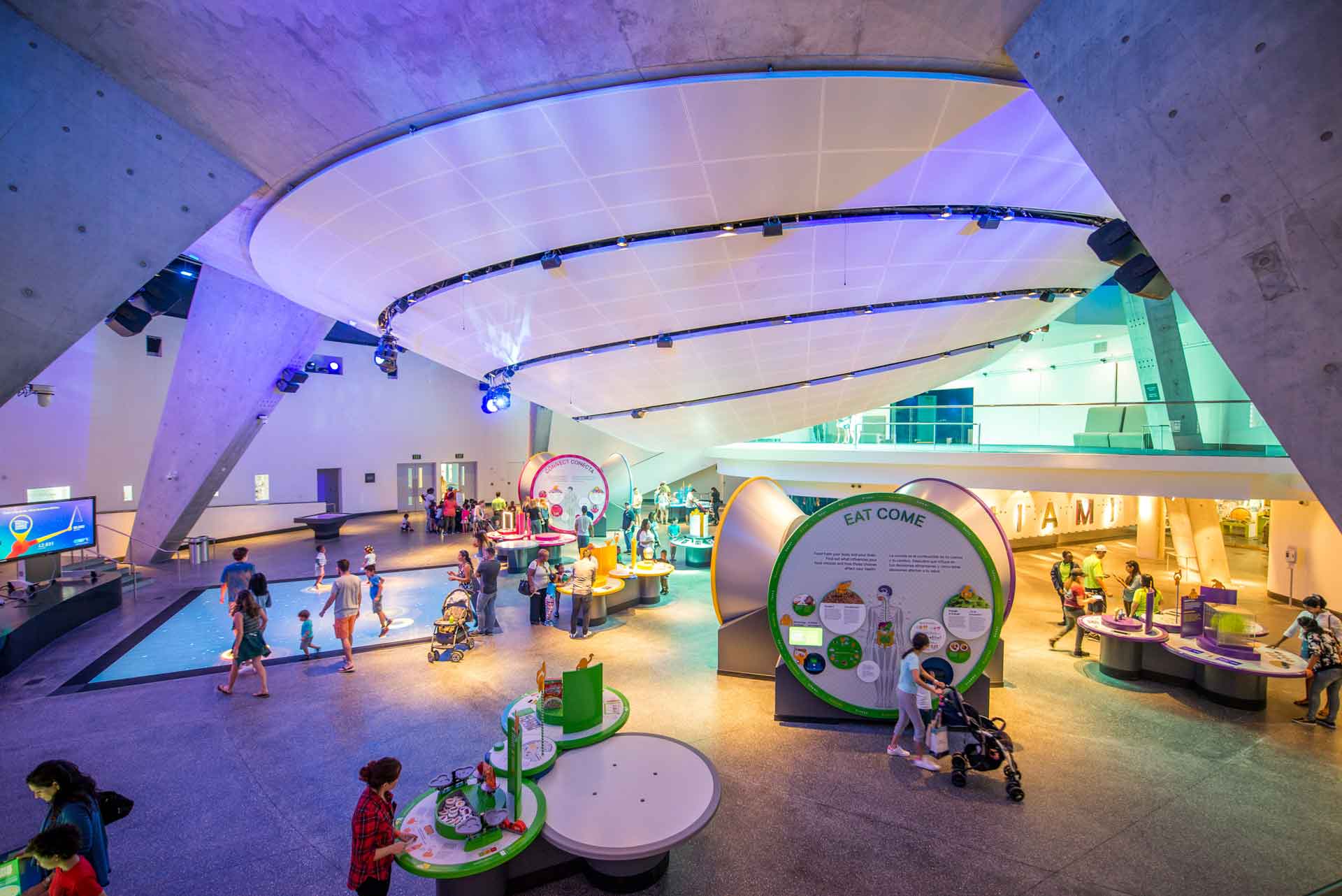 A modern building interior features a curved, white ceiling extending over an interactive exhibition space where visitors engage with circular green and white educational displays about nutrition.