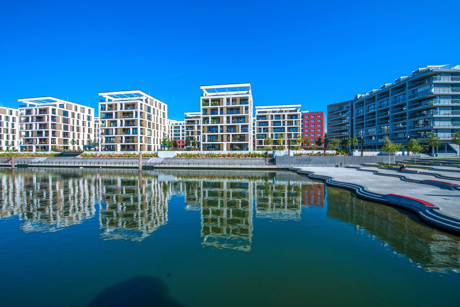 A contemporary urban riverside development with several white structures adorned with balconies and red highlights, mirrored in the tranquil waters of a neighboring river or canal beneath a clear blue sky.