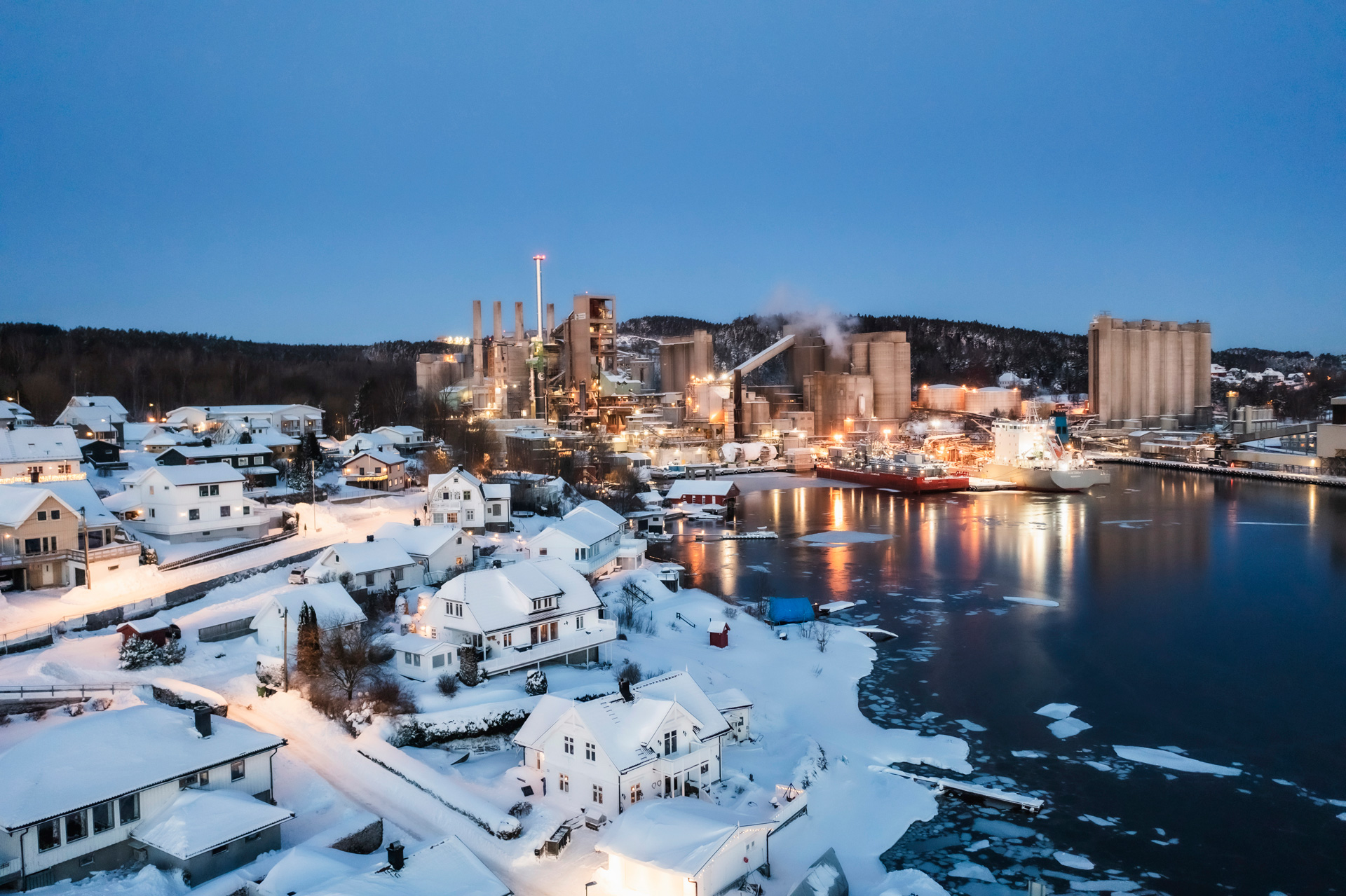 Snow-covered town by the water during twilight. In the center, there’s a cement plant with smokestacks emitting vapor. Ships are docked at the harbor nearby. Residential houses with steep roofs dot the area, and a large storage facility stands on the right side. The presence of snow and ice in the water suggests cold temperatures.