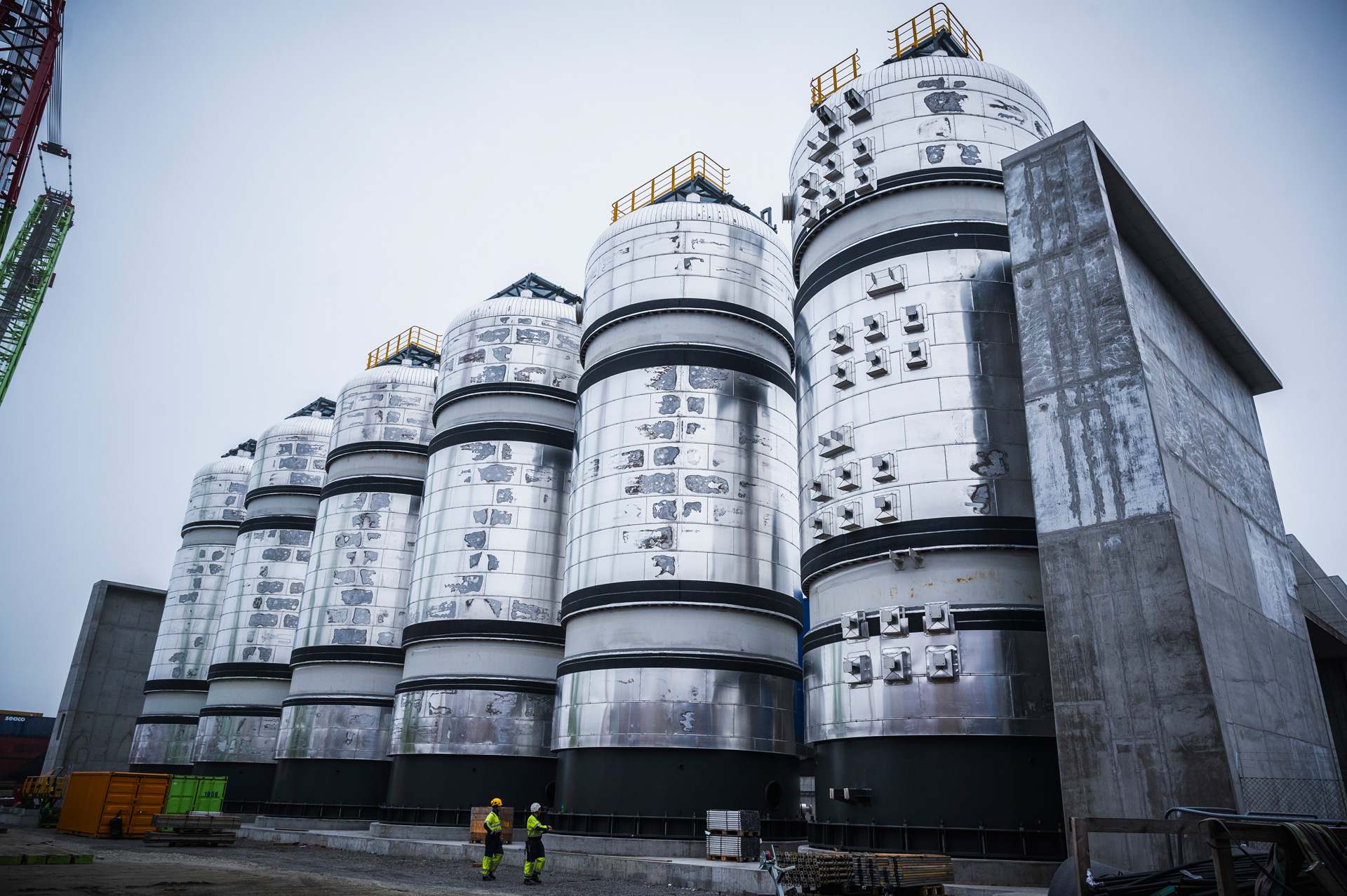 Series of large cylindrical structures with conical tops, resembling industrial silos. These metal structures have visible access points, ladders, and safety railings. Despite their size, they appear even more massive when compared to the two individuals standing at their base, both wearing safety gear. The overcast sky suggests an industrial setting on a cloudy day.