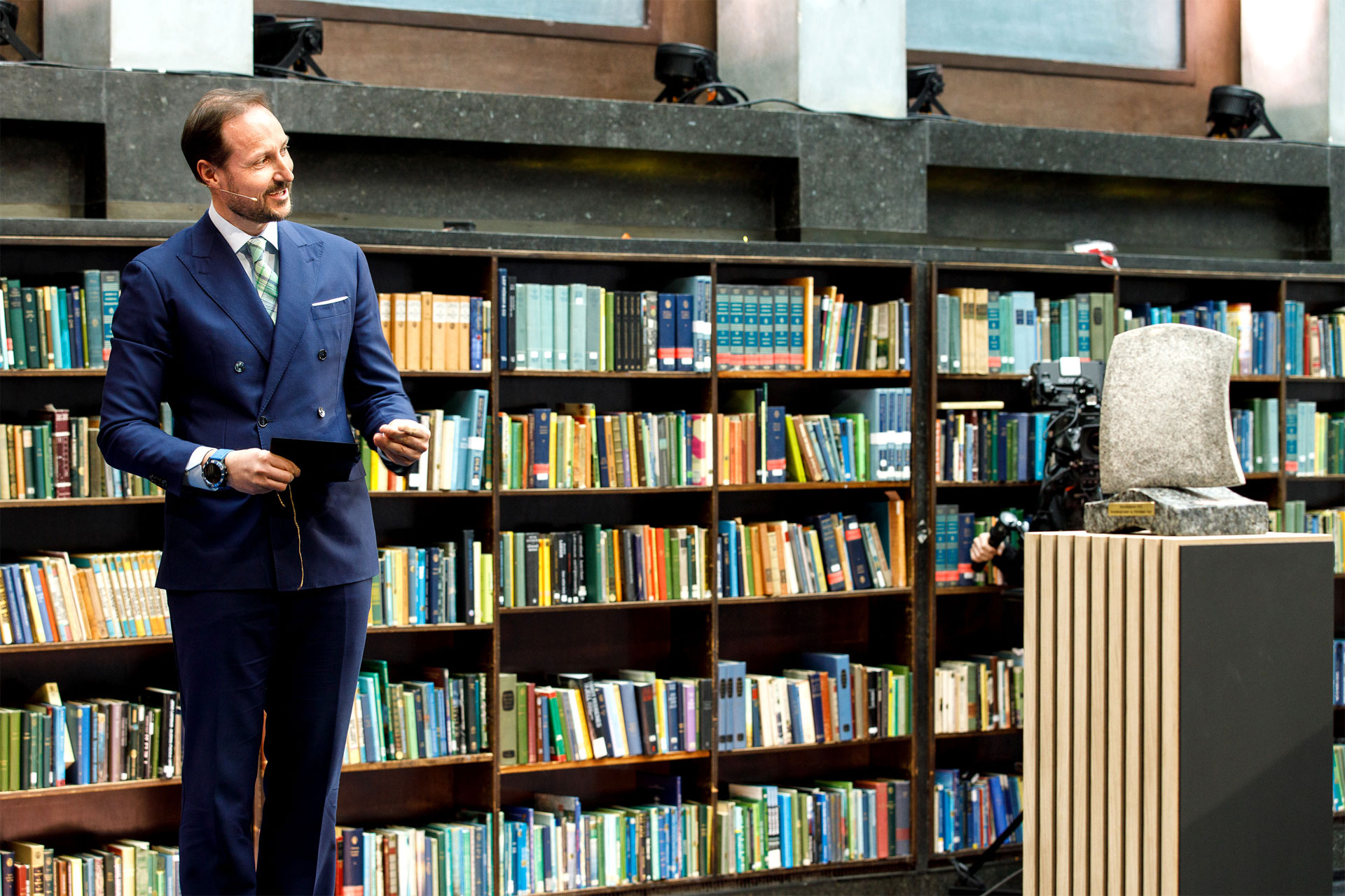 A person in a blue suit standing in front of a bookshelf filled with colourful books, with an abstract sculpture on a pedestal to the right
