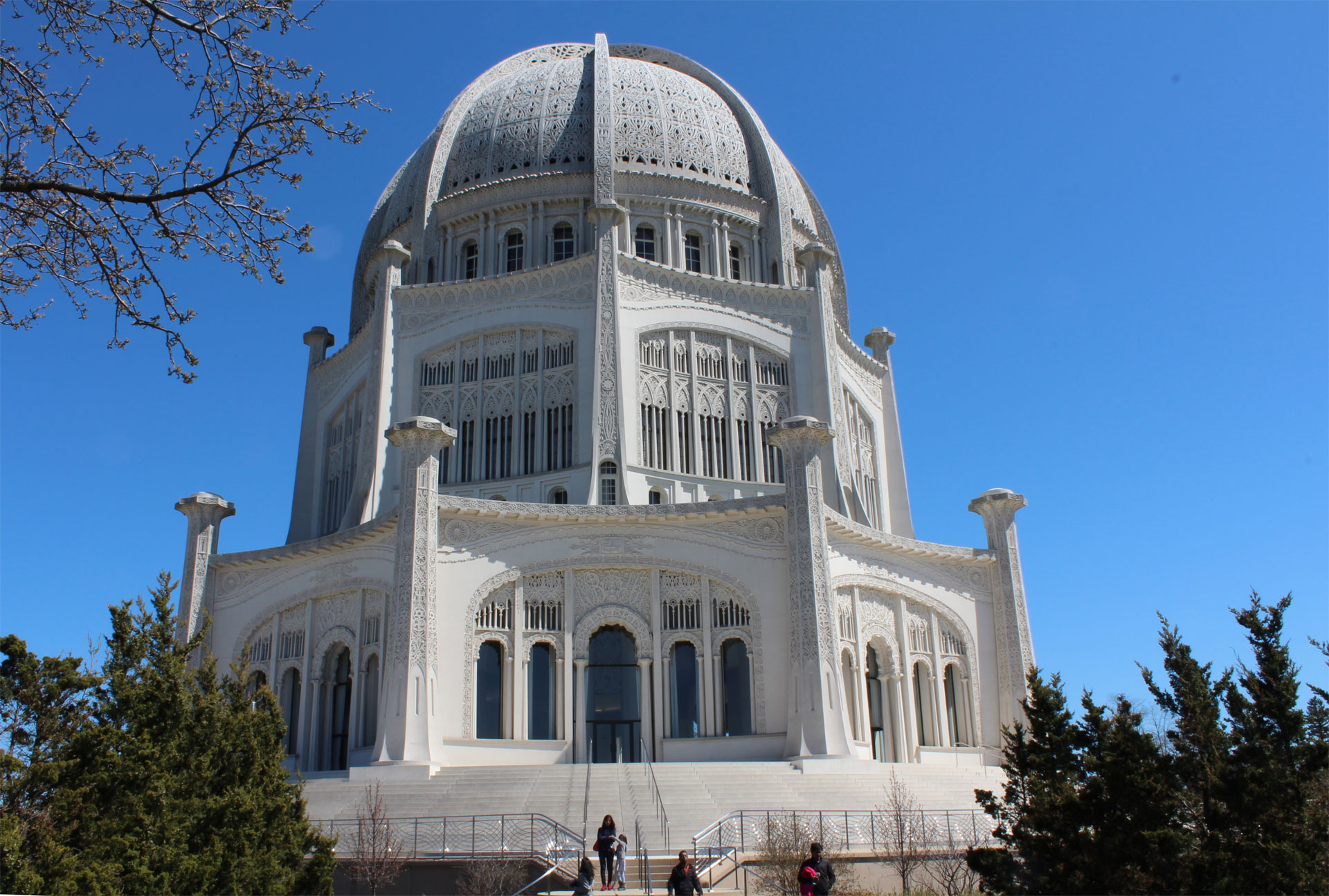 A majestic white building with a large dome and multiple arches, set against a clear blue sky, surrounded by greenery