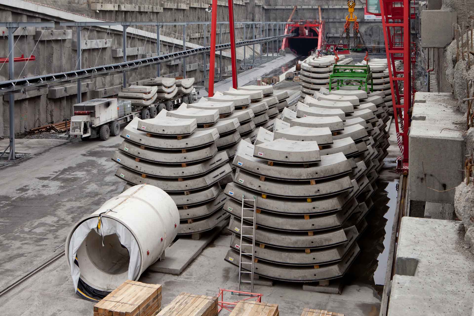 Ein Baustellenbild zeigt gestapelte Segmente eines Tunnelfuttersystems vor dem Hintergrund einer ausgehobenen Fläche mit einer roten Tunnelbohrmaschine im Hintergrund