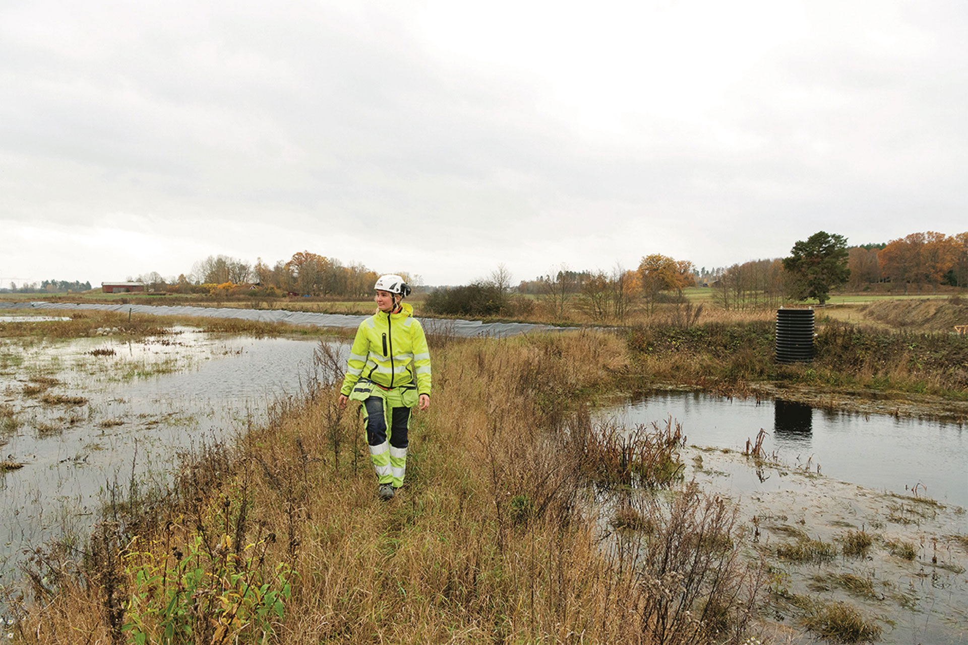 A person in protective equipment and a hard hat standing by a pond in a grassy field, with overcast skies above