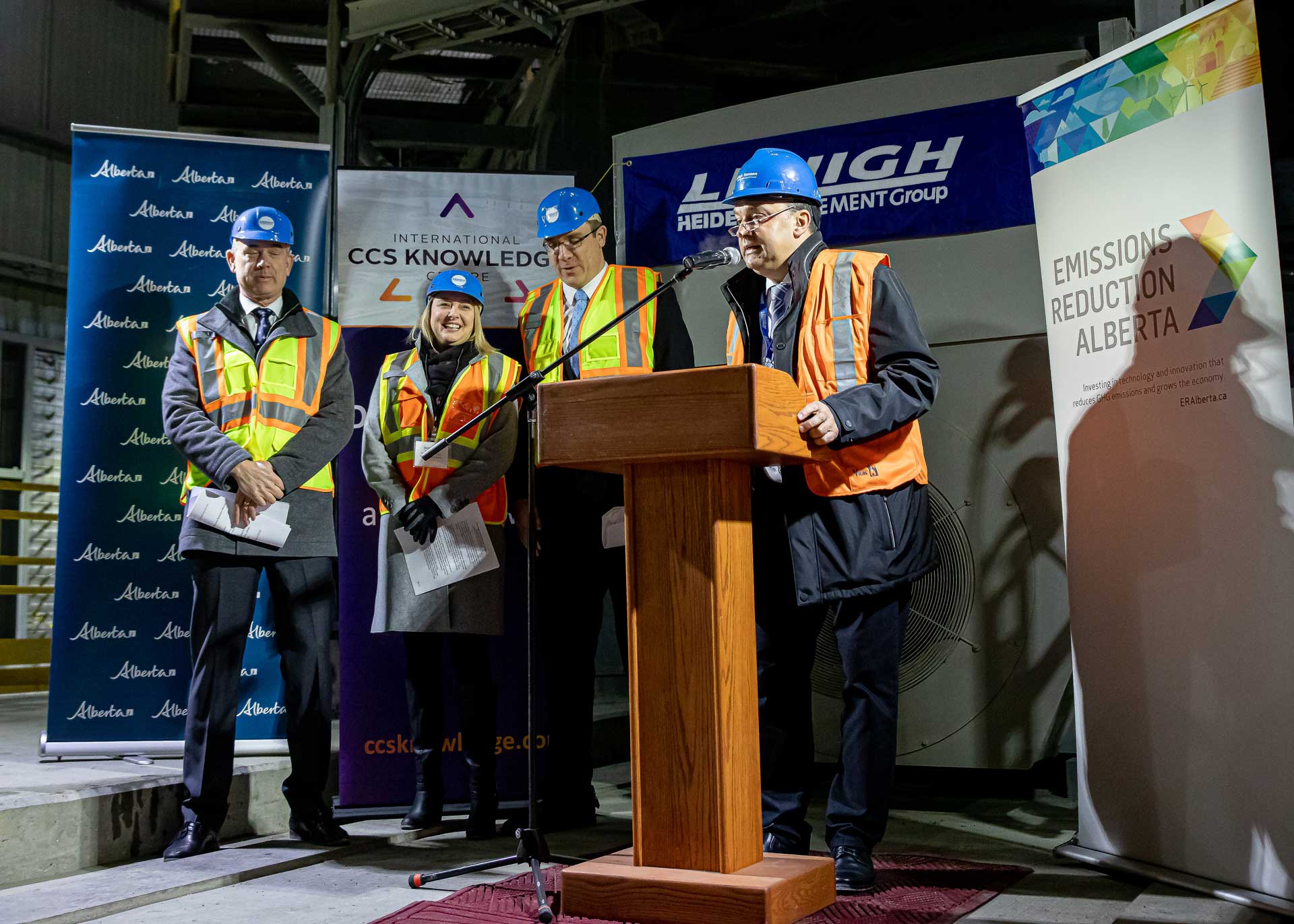 The image shows four individuals standing behind a podium in an industrial environment, their faces not visible. Banners with the logos “CCS KNOWLEDGE,” “HEIDELBERGCEMENT,” and “EMISSION REDUCTION ALBERTA” are displayed in the background.