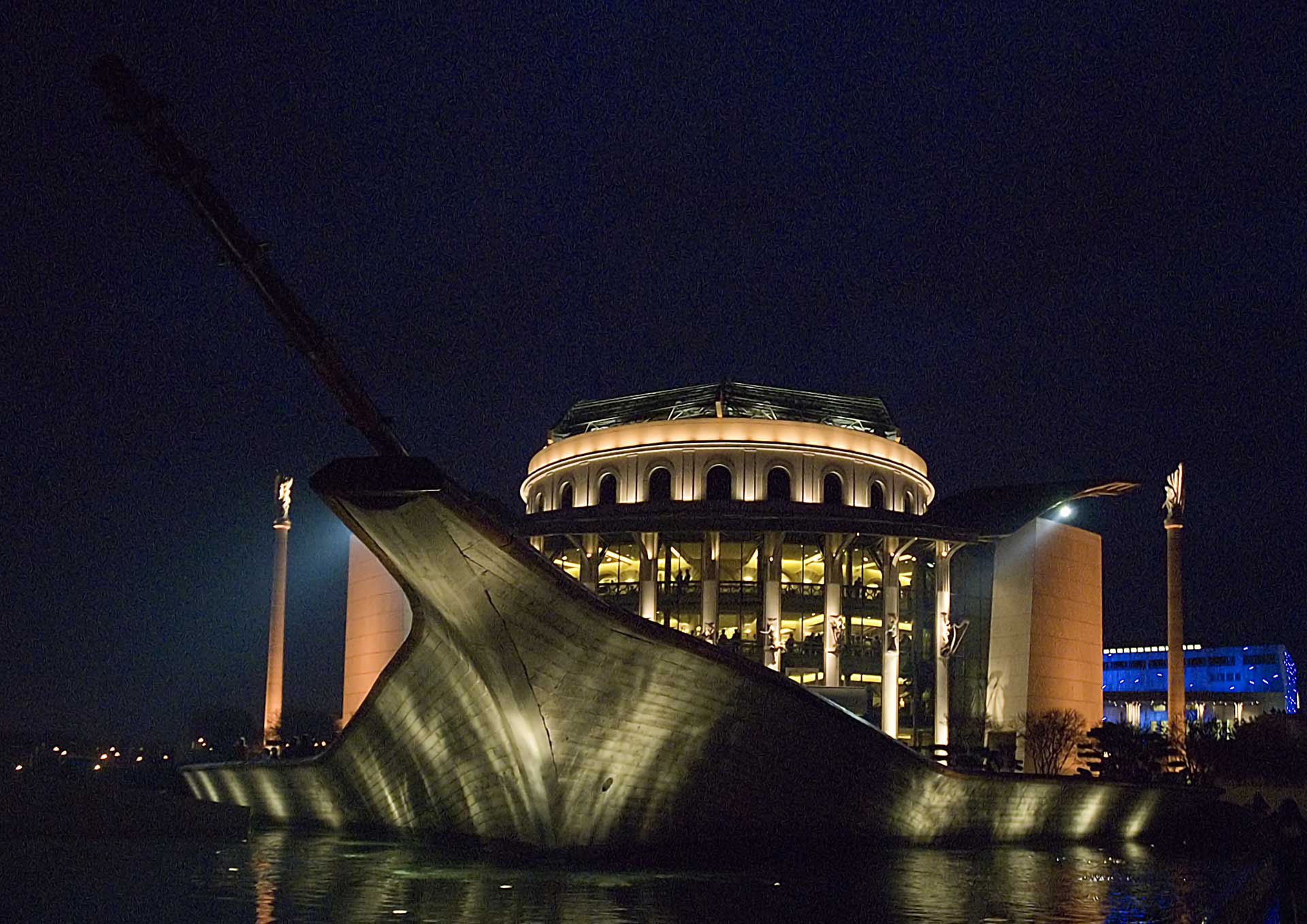 Palast der Künste, Ein nächtliches Gebäude, das einem Schiff ähnelt, mit einem Bug, der sich nach oben und außen wölbt, als ob es aus dem Wasser auftaucht, beleuchtet durch innere Lichter, die runde Fenster und eine kuppelartige Spitze hervorheben.