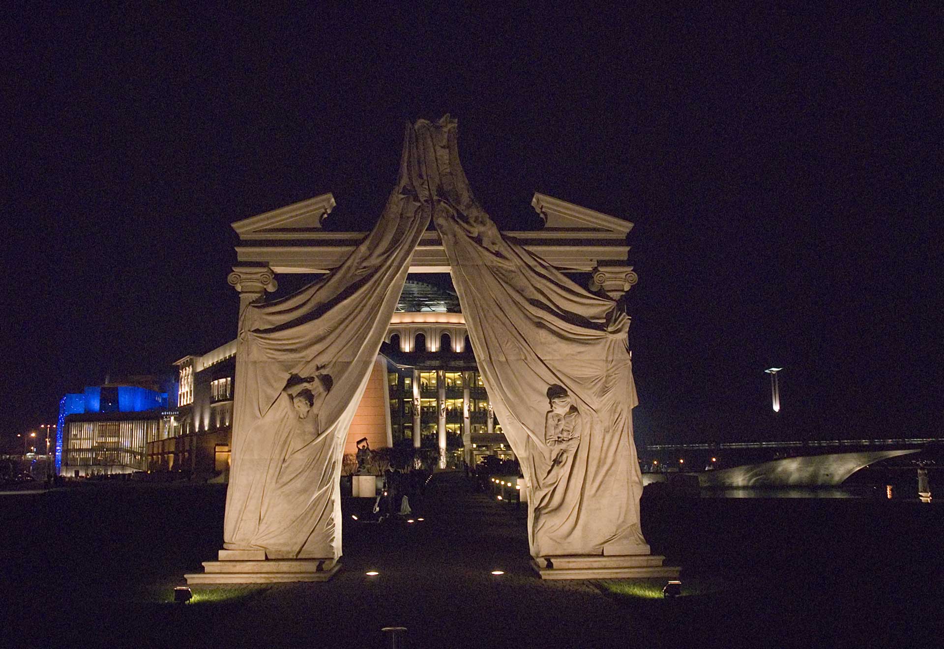 An illuminated classical arch, draped with a white cloth featuring bas-relief sculptures, stands in contrast to a modern building with blue lights and glass facades in the background.