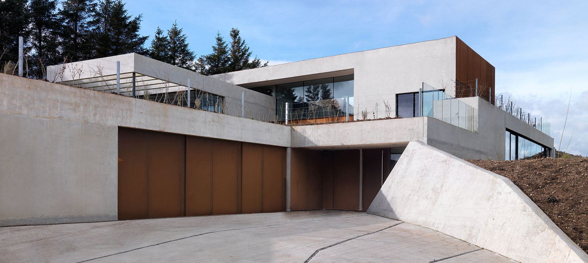 Ein modernes Wohnhaus mit geometrischem Design, das Beton und Holz kombiniert, mit großen Fenstern und einem Flachdach, unter einem teilweise bewölkten Himmel