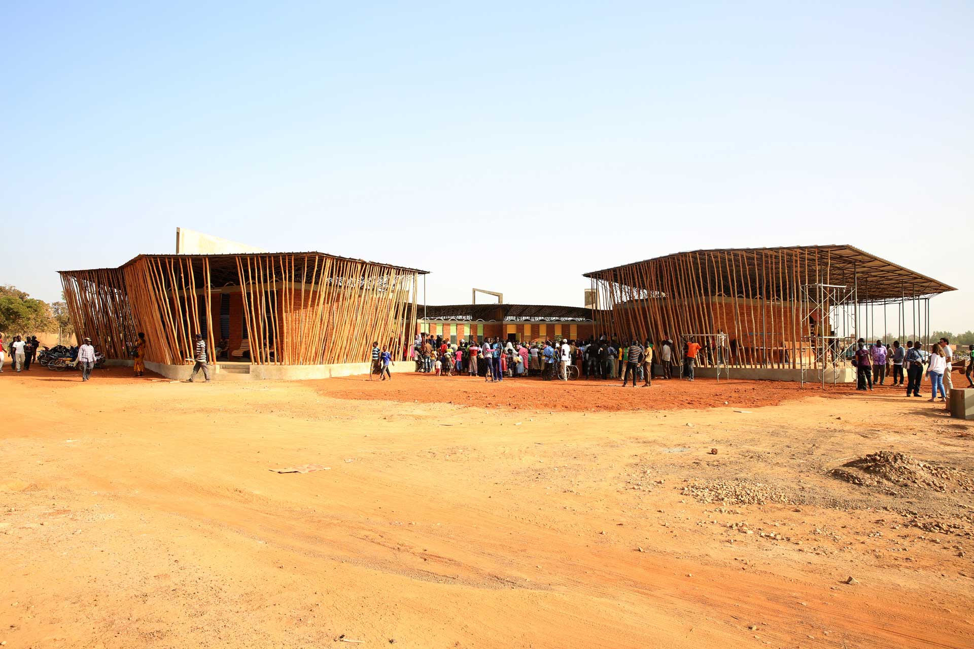 group of individuals standing in front of two large wooden and bamboo structures that are either under construction or designed in an open framework style, with a natural landscape in the background