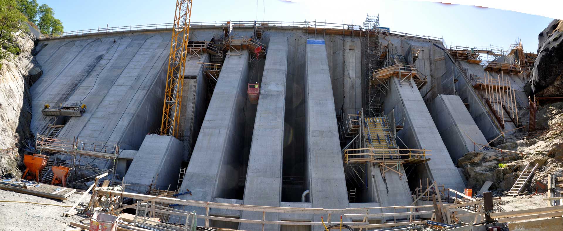 Ein im Bau befindlicher großer Betondamm mit zahlreichen Stützstrukturen und Baumaschinen, umgeben von felsigem Terrain unter einem klaren Himmel.