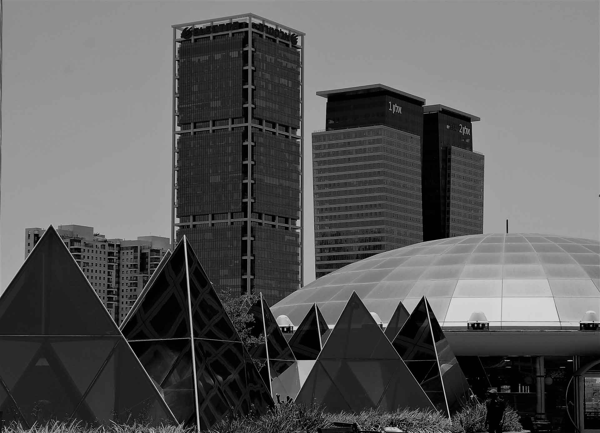 A cityscape showcases modern architecture with pyramid-like structures in the foreground and towering buildings behind. The tallest building features a grid-patterned facade, flanked by two buildings with distinct logos, all under a monochrome sky.