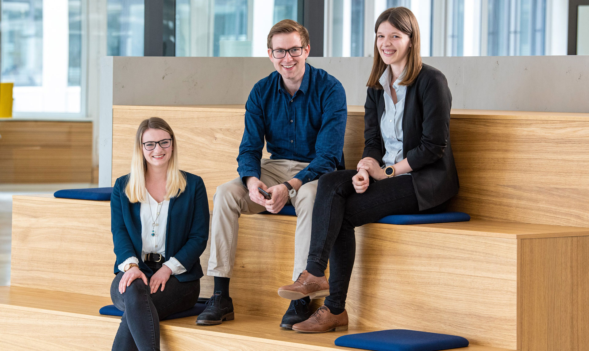 Three individuals are seated on a wooden bench inside a modern building, their faces obscured for privacy. One is wearing a light blue blouse and dark trousers, another a blue shirt and jeans, and the third is dressed in a black blazer and skirt.