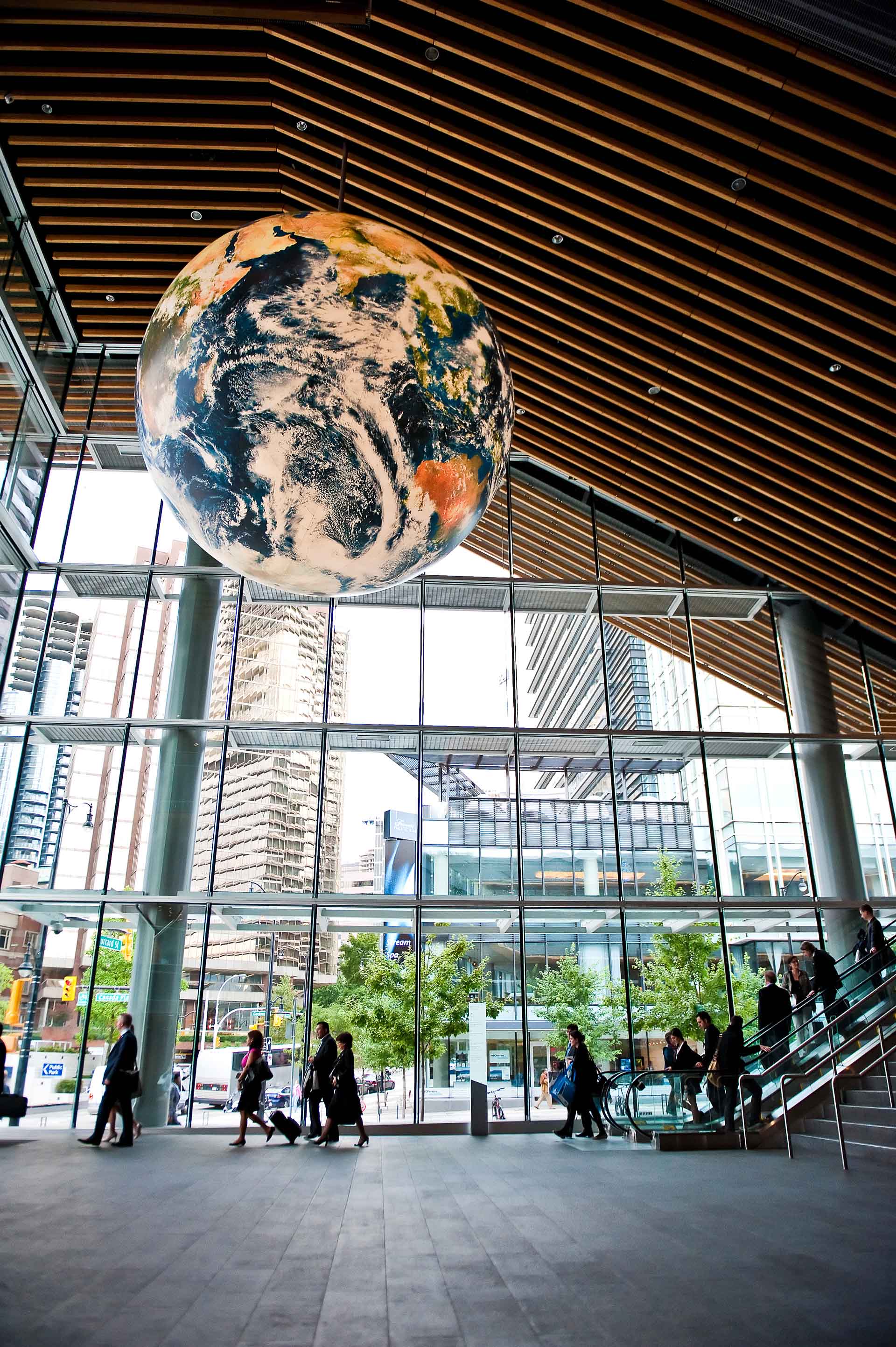 Ein markanter Globus hängt im Zentrum eines modernen öffentlichen Raumes, umgeben von Menschen und Treppen, und symbolisiert einen Schnittpunkt von Kultur und Lernen.