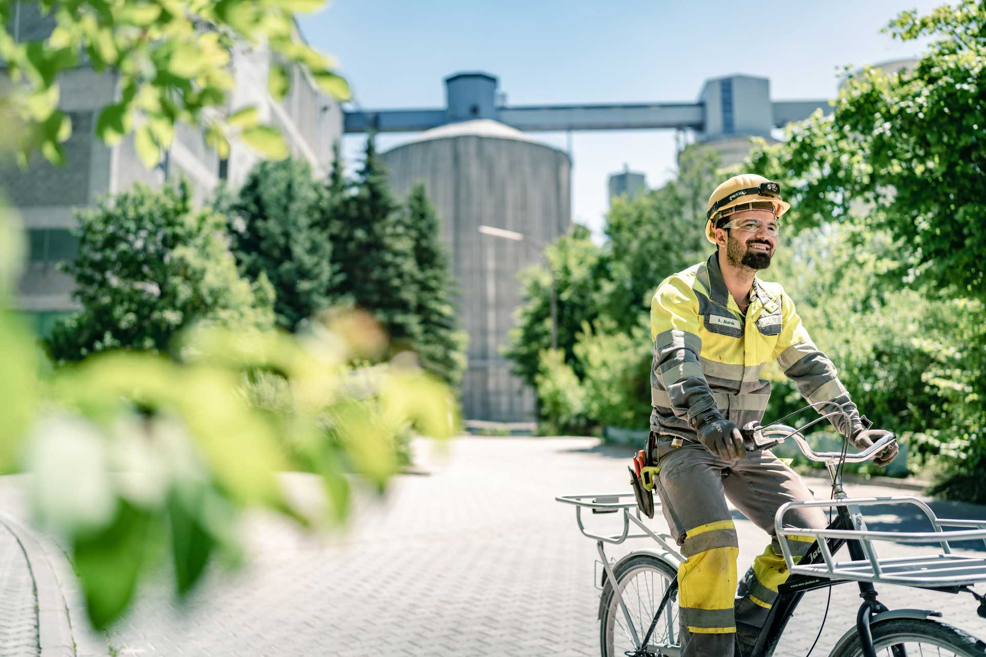 A person in yellow and black work attire with a helmet rides a bicycle. Large industrial silos and lush green trees are visible under a clear blue sky in the background.