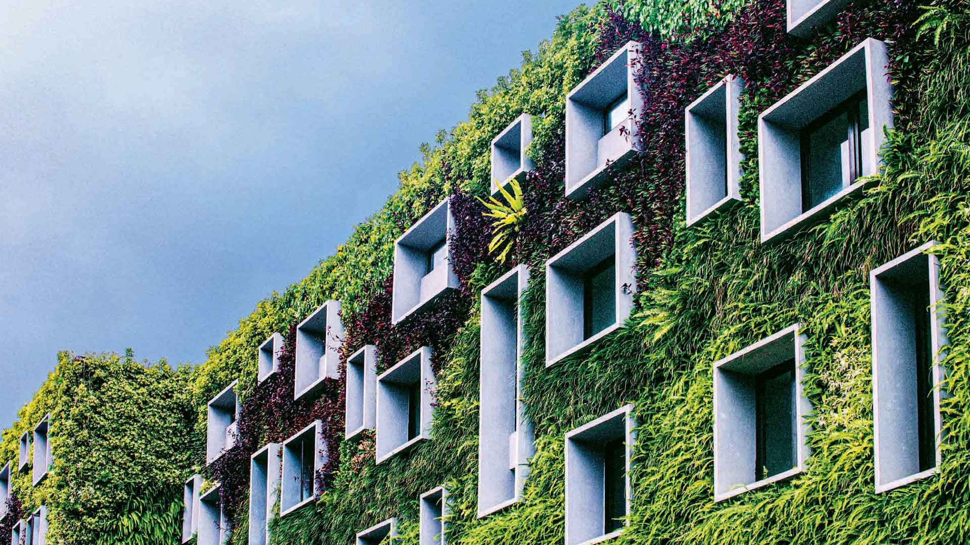 A contemporary building facade is enveloped in a vertical garden of lush greenery, exemplifying sustainable architecture that blends nature into the urban landscape. The design features protruding rectangular sections, adding dimension and architectural intrigue to the edifice.