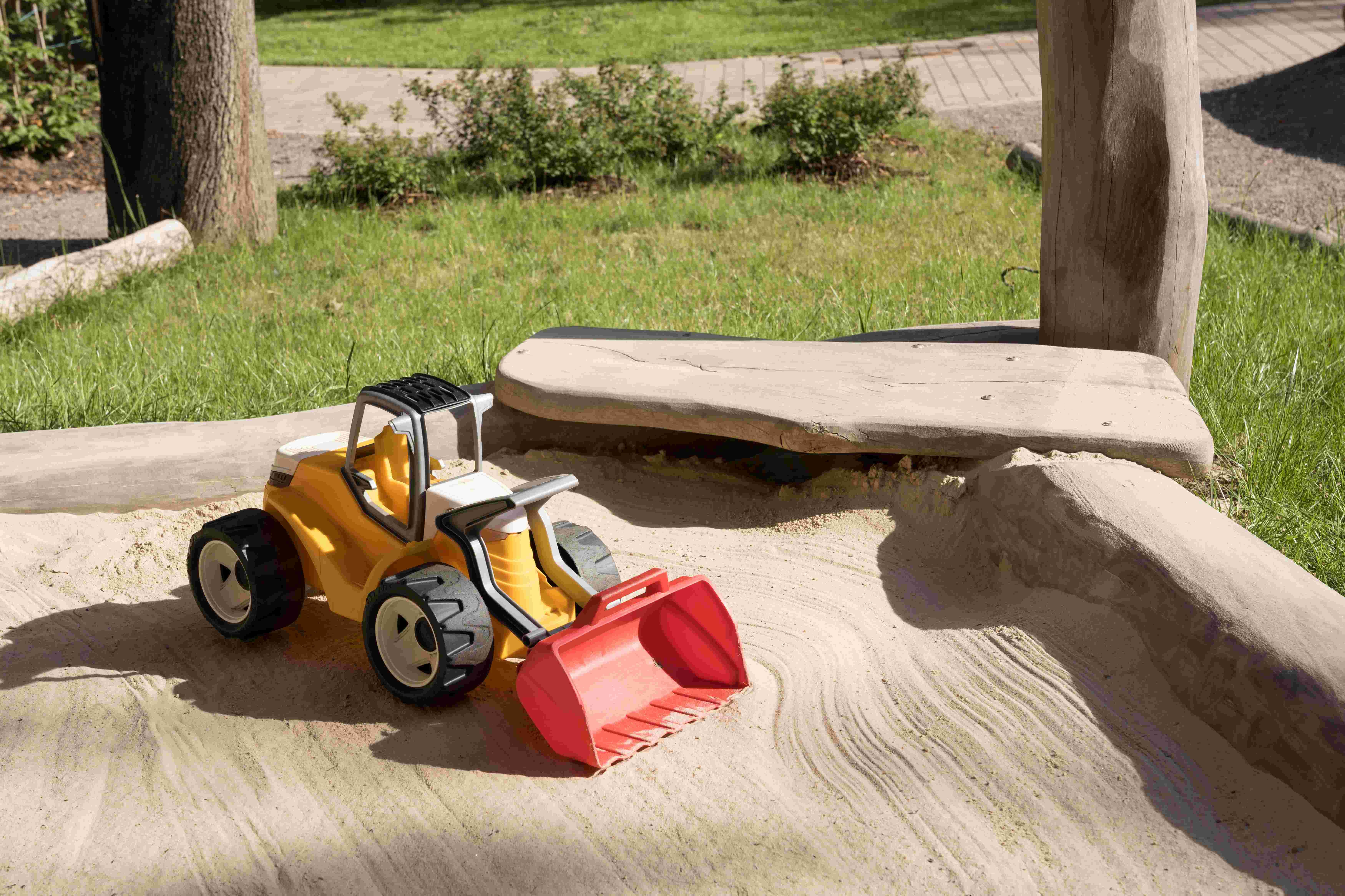 Ein Spielzeug-Bulldozer auf einer sandigen Fläche, mit einem Betonrandstein und Gras im Hintergrund. Der Bulldozer ist so positioniert, als ob er bereit wäre, den Sand zu schieben oder zu bewegen.