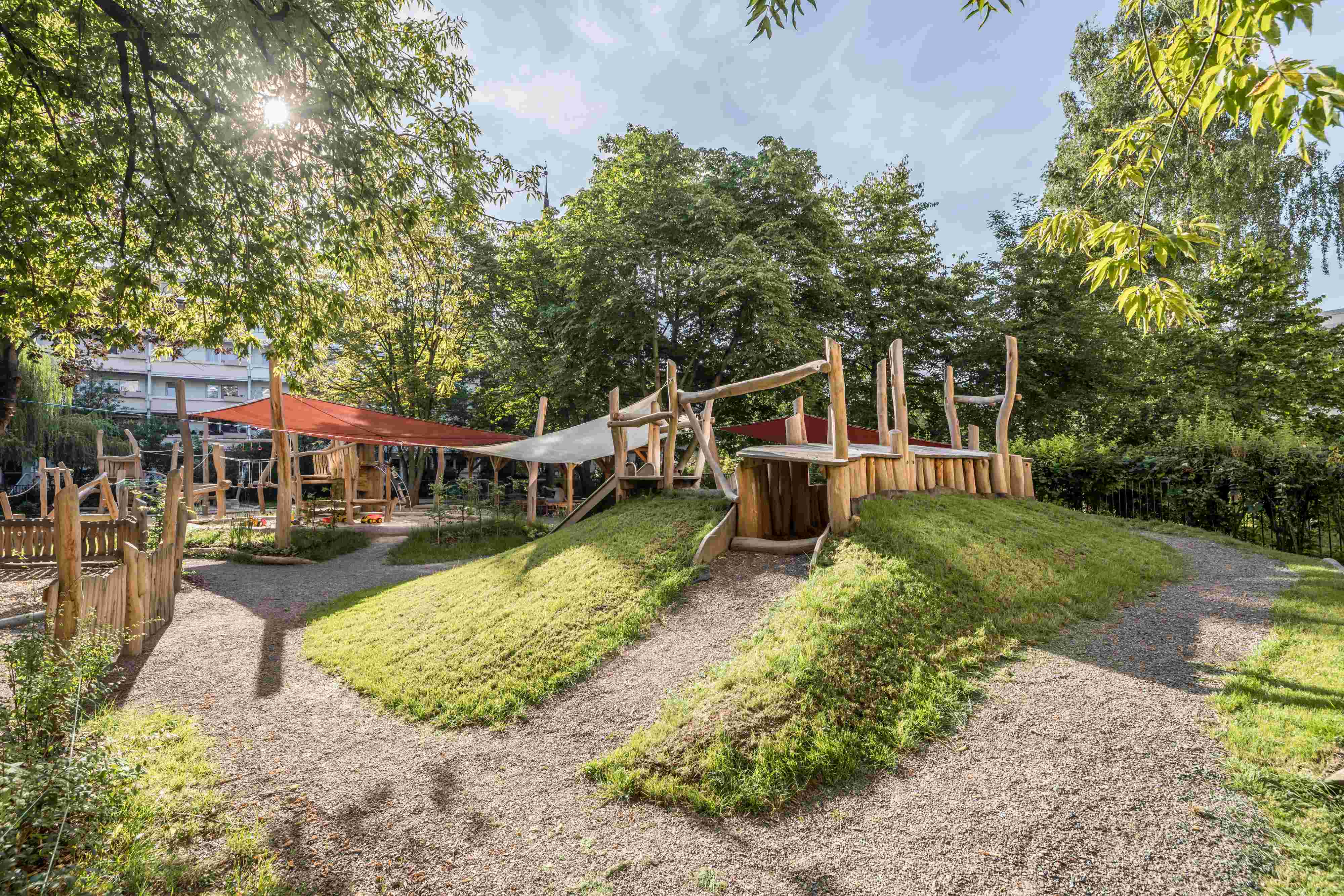 An outdoor playground set in a lush green area with trees and sunlight filtering through the foliage. The playground includes wooden structures such as a playhouse, bridges, and platforms, as well as grassy mounds.