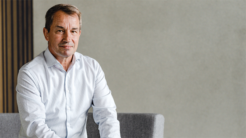 Dr Wolfgang Dienemann seated on a grey sofa wearing a white button-up shirt. The background is blurred with neutral tones.