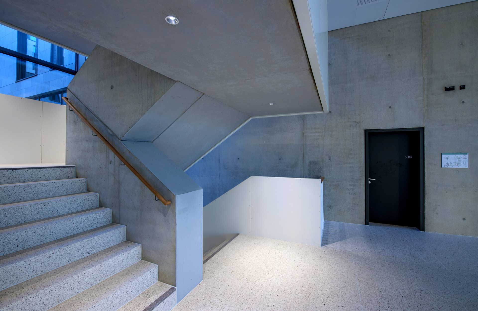 A contemporary interior with a concrete staircase and metal handrail on the right leading to another level. Below the stairs is a black door; the space is illuminated by natural light from the left.