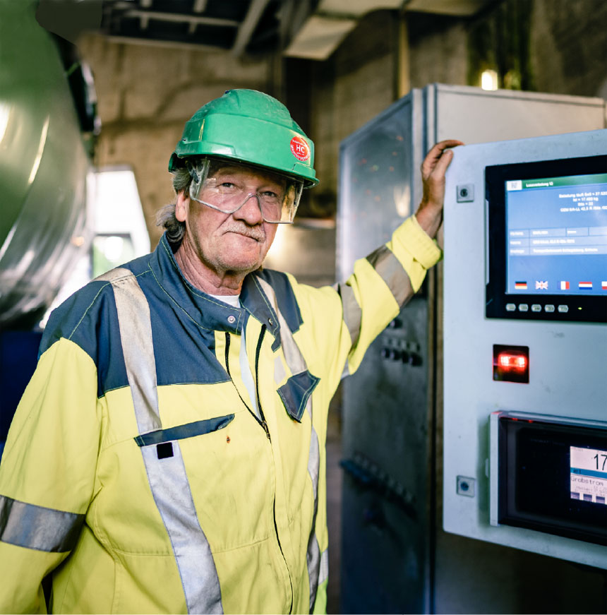 An industrial worker in a high-visibility jacket and green hard hat operating a machine with a digital screen.