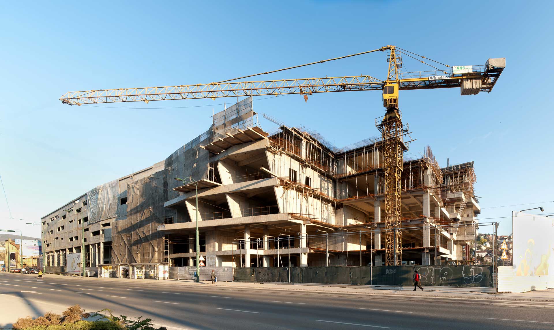 Eine große Baustelle bei Tageslicht. Ein mehrstöckiges Gebäude im Bau mit sichtbarem Gerüst und unvollständigen Strukturelementen. Ein hoher gelber Kran ragt über das Gebäude, was auf laufende Arbeiten hinweist.