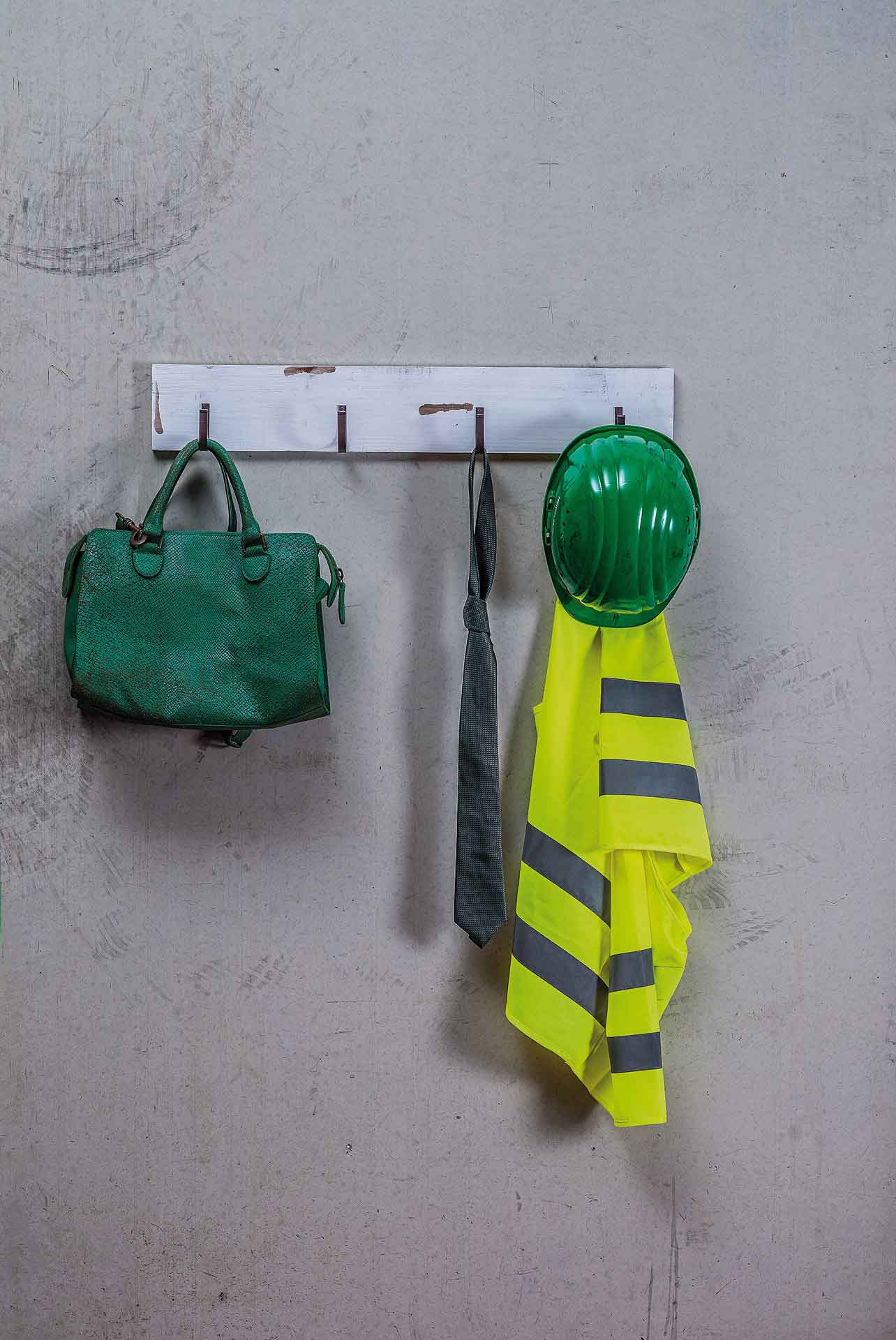 A green satchel, grey tie, upside-down hard hat and high-visibility vest on wall hooks against a textured grey concrete background.