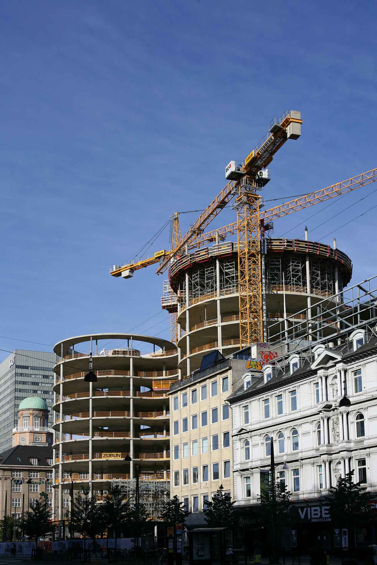 Eine Baustelle mit einem großen gelben Kran, der über einem teilweise errichteten mehrstöckigen Gebäude ragt. Im Vordergrund befinden sich traditionelle Gebäude mit klassischer Architektur, was darauf hinweist, dass die Baustelle in einem städtischen Gebiet liegt, das neue Entwicklungen mit alten Stadtstrukturen verbindet.