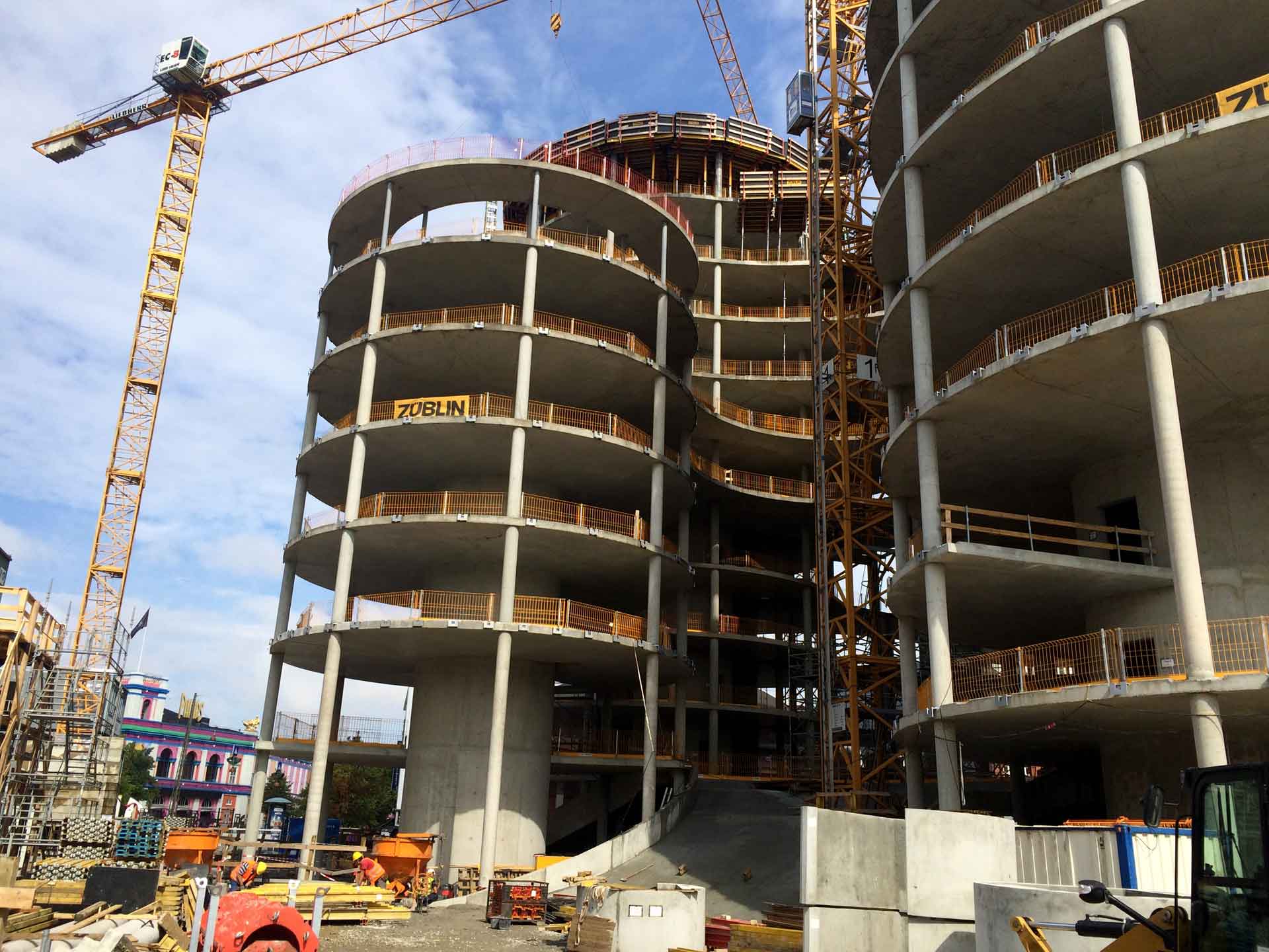 Eine Baustelle mit einem mehrstöckigen Gebäude im Bau. Das Gebäude hat ein geschwungenes Design mit mehreren sichtbaren Stockwerken.